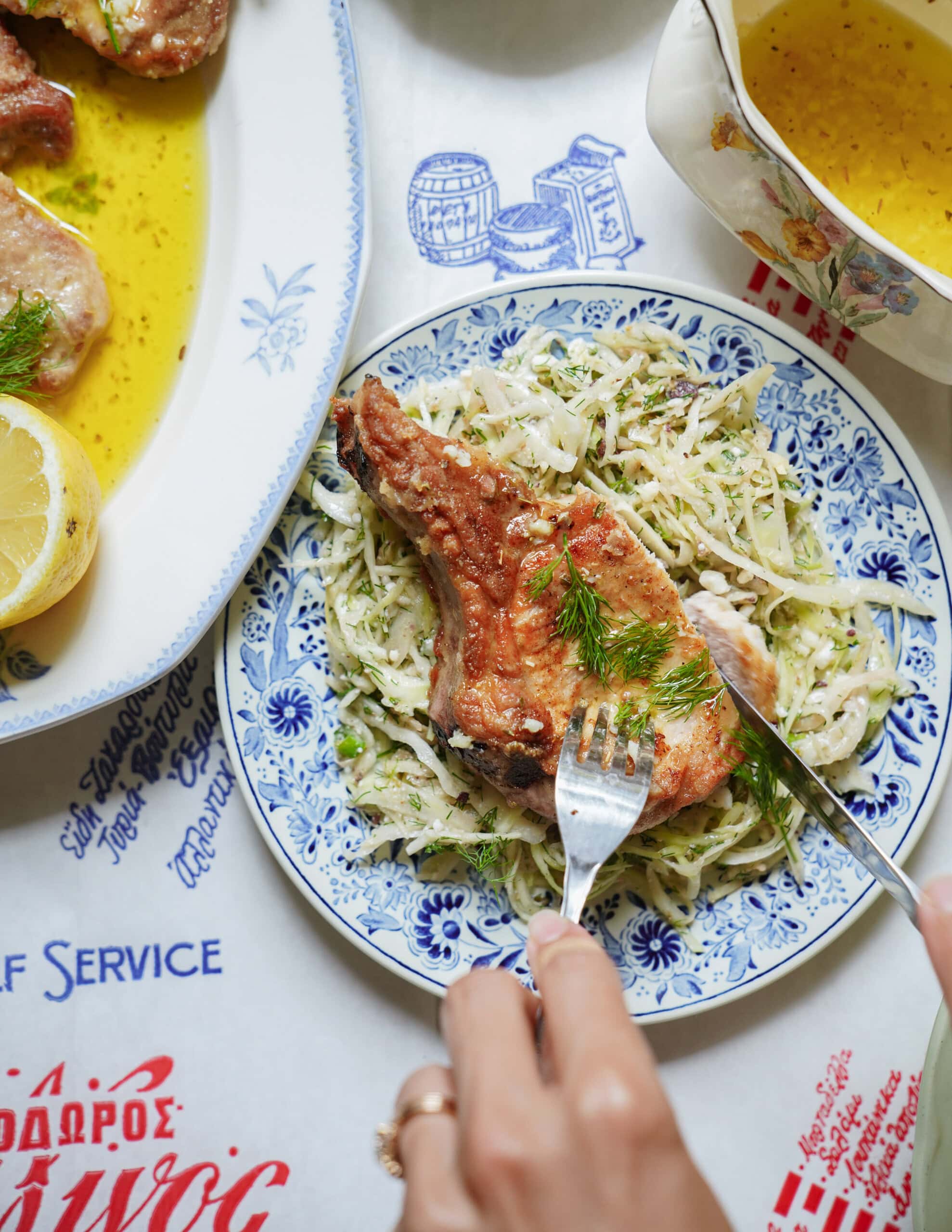 A hand cuts into a crispy pork chop served on a bed of shredded cabbage salad, garnished with fresh dill, on a blue and white plate. A lemon wedge and sauce are visible nearby on the table.
