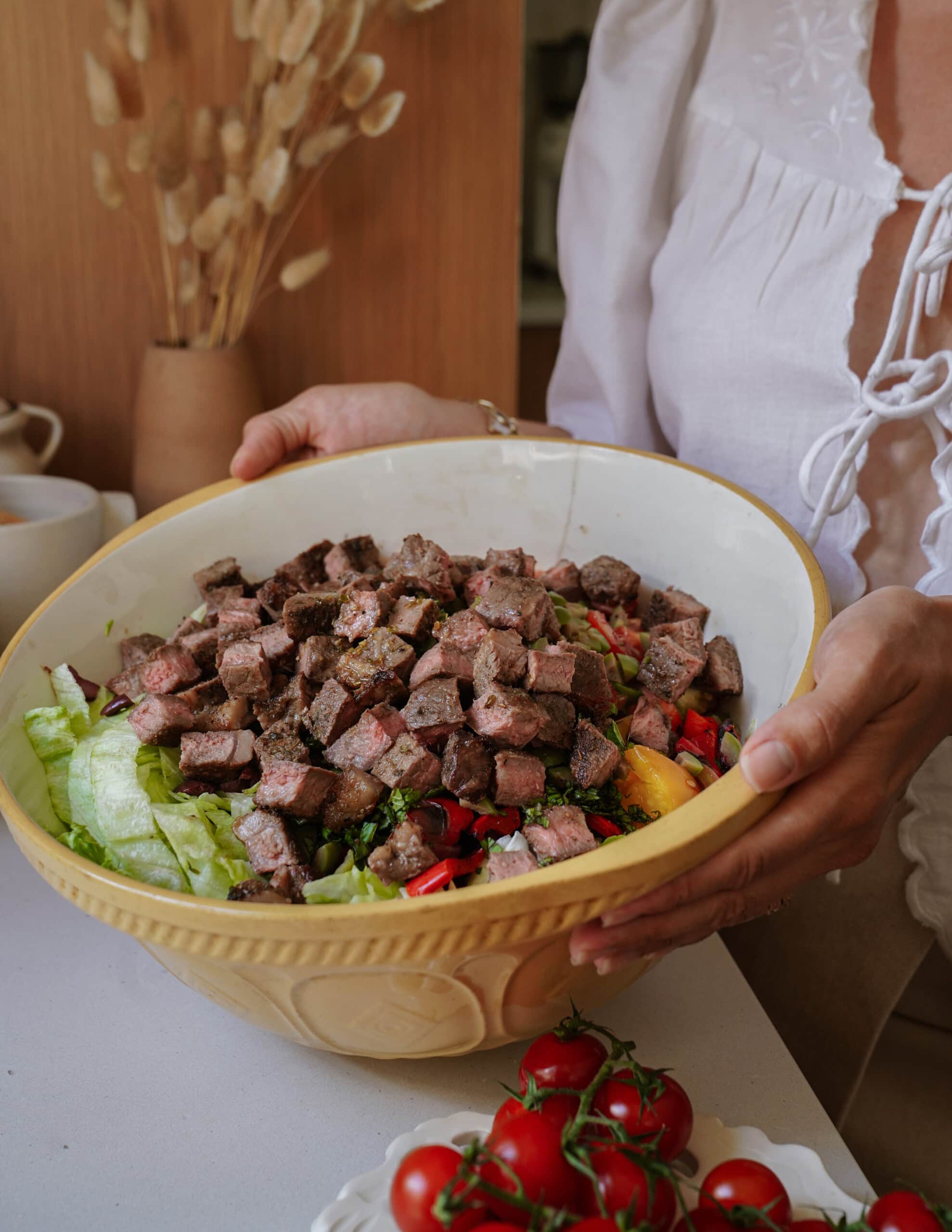 A person in a white blouse holds a large yellow bowl filled with salad topped with chunks of cooked steak. Fresh tomatoes on the vine sit on the counter nearby.