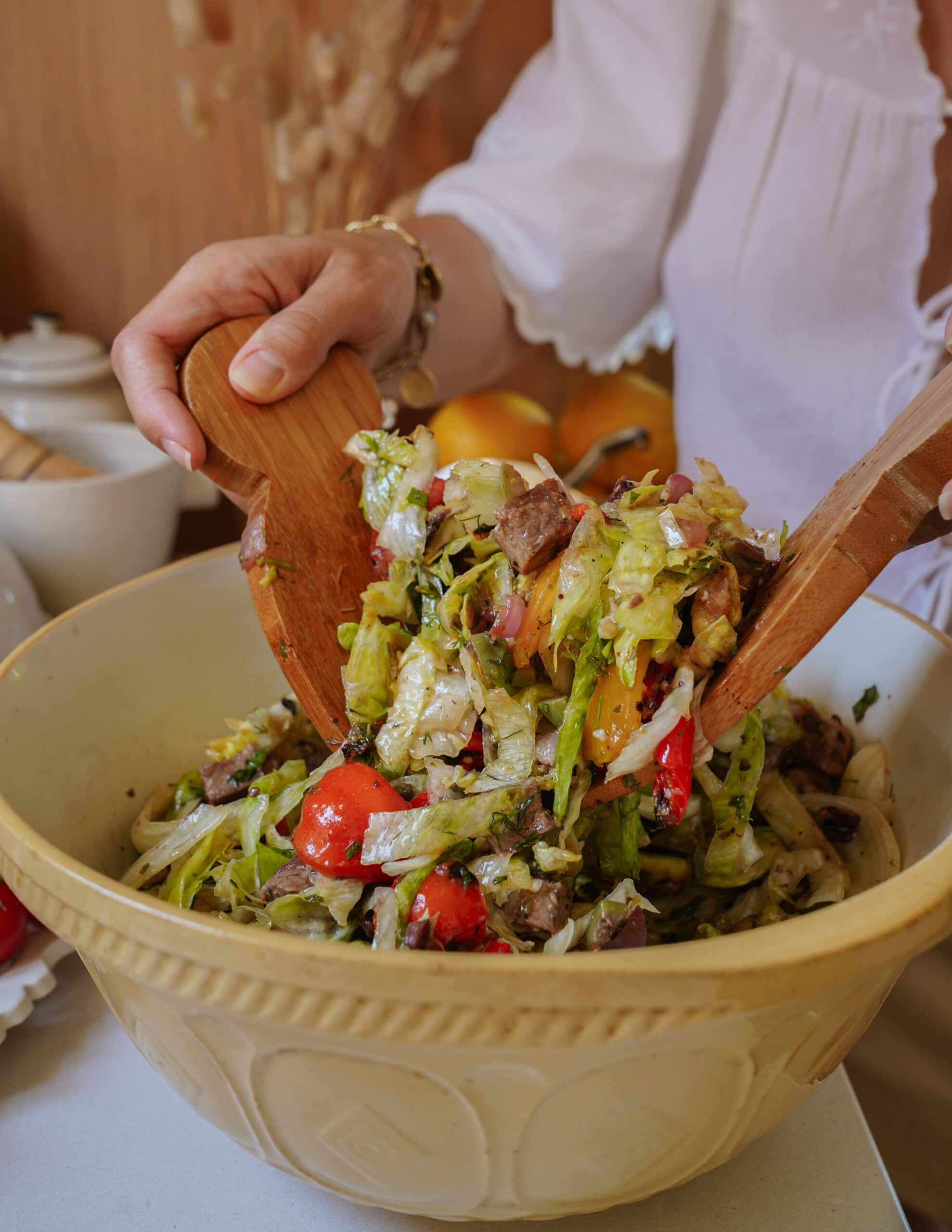 A person in a white shirt tosses a fresh salad with wooden utensils in a large beige bowl. The salad contains lettuce, tomatoes, onions, and other vegetables.