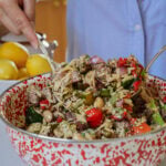 A person in a blue shirt mixes a colorful orzo salad with vegetables, beans, and herbs in a large red and white speckled bowl. Lemons sit on the counter in the background.