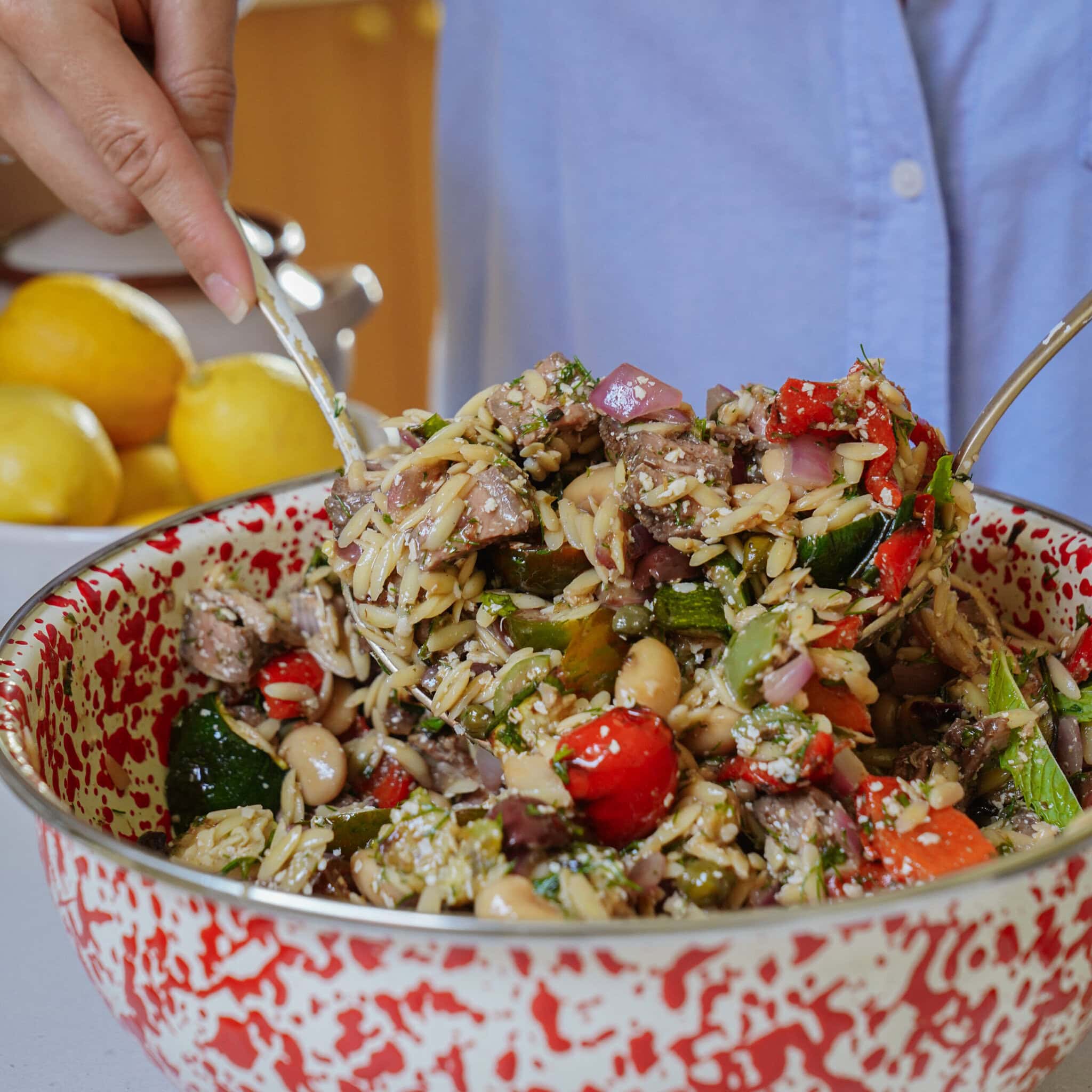 A person in a blue shirt mixes a colorful orzo salad with vegetables, beans, and herbs in a large red and white speckled bowl. Lemons sit on the counter in the background.