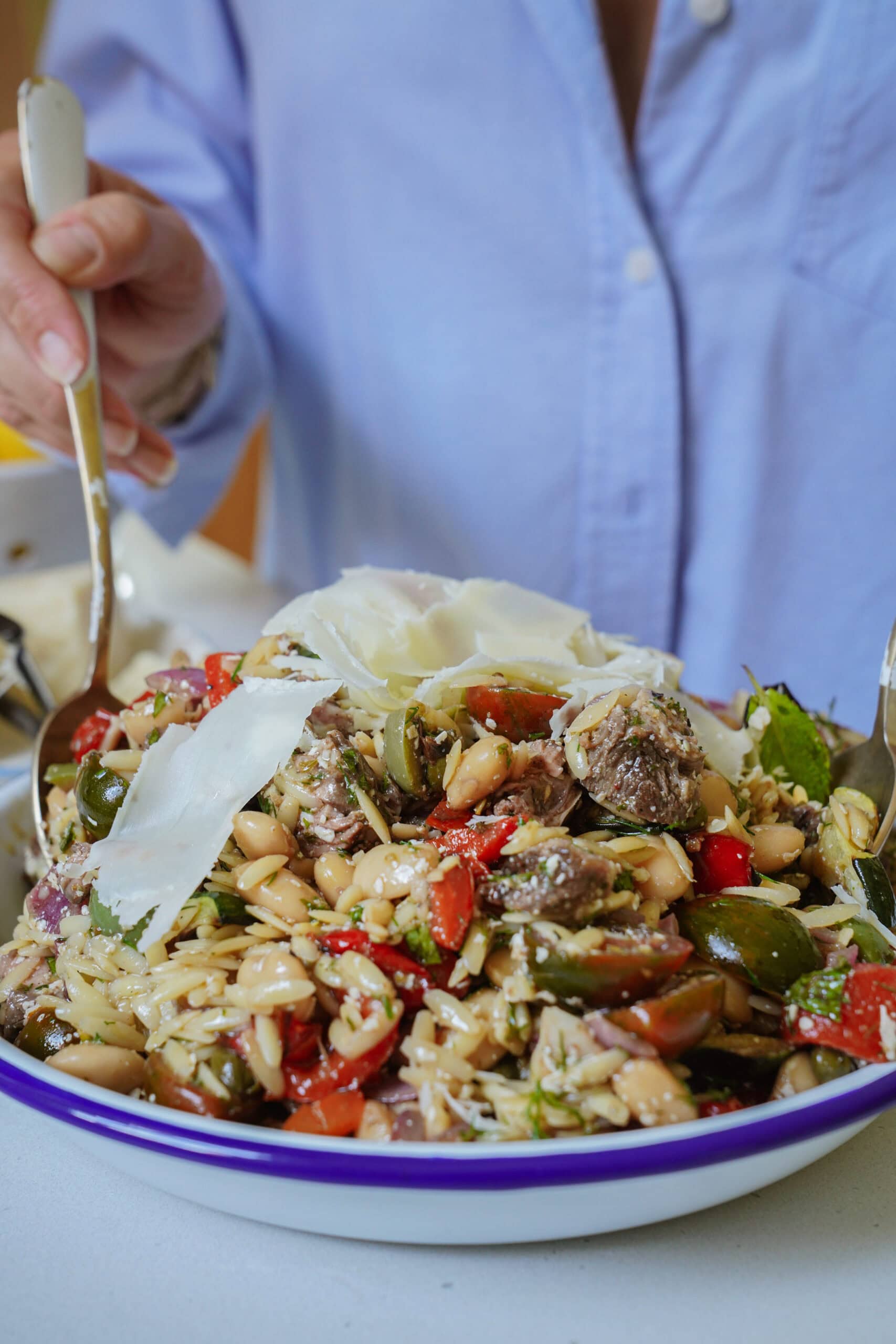 A person in a blue shirt holds a fork and spoon, serving a colorful salad with orzo, beans, tomatoes, greens, and large shavings of cheese on top, all in a white bowl with a blue rim.