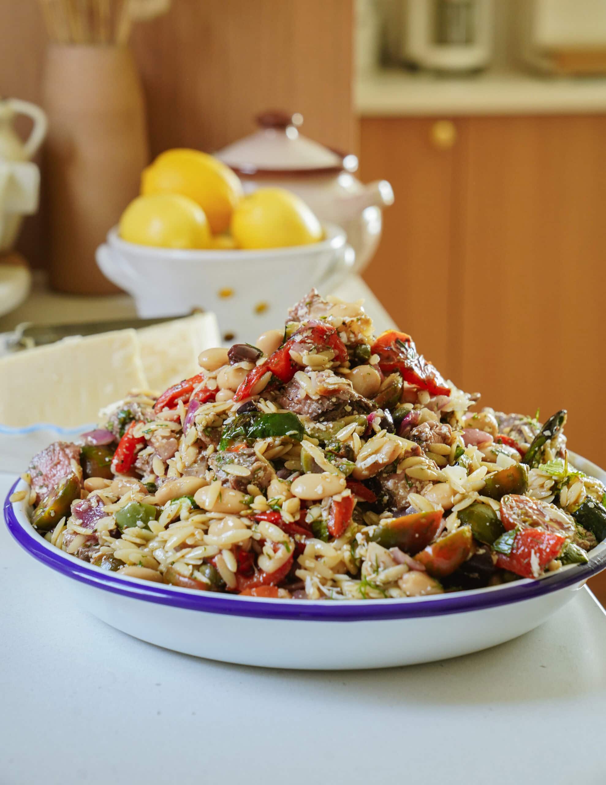 A bowl of colorful orzo pasta salad with vegetables, beans, and tuna sits on a kitchen counter. In the background, there is a bowl of lemons and a block of cheese.