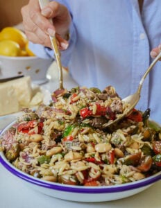 A person in a light blue shirt uses two forks to serve a colorful orzo salad with beans, tomatoes, olives, and herbs from a large, shallow bowl. Lemons and a block of cheese are visible in the background.