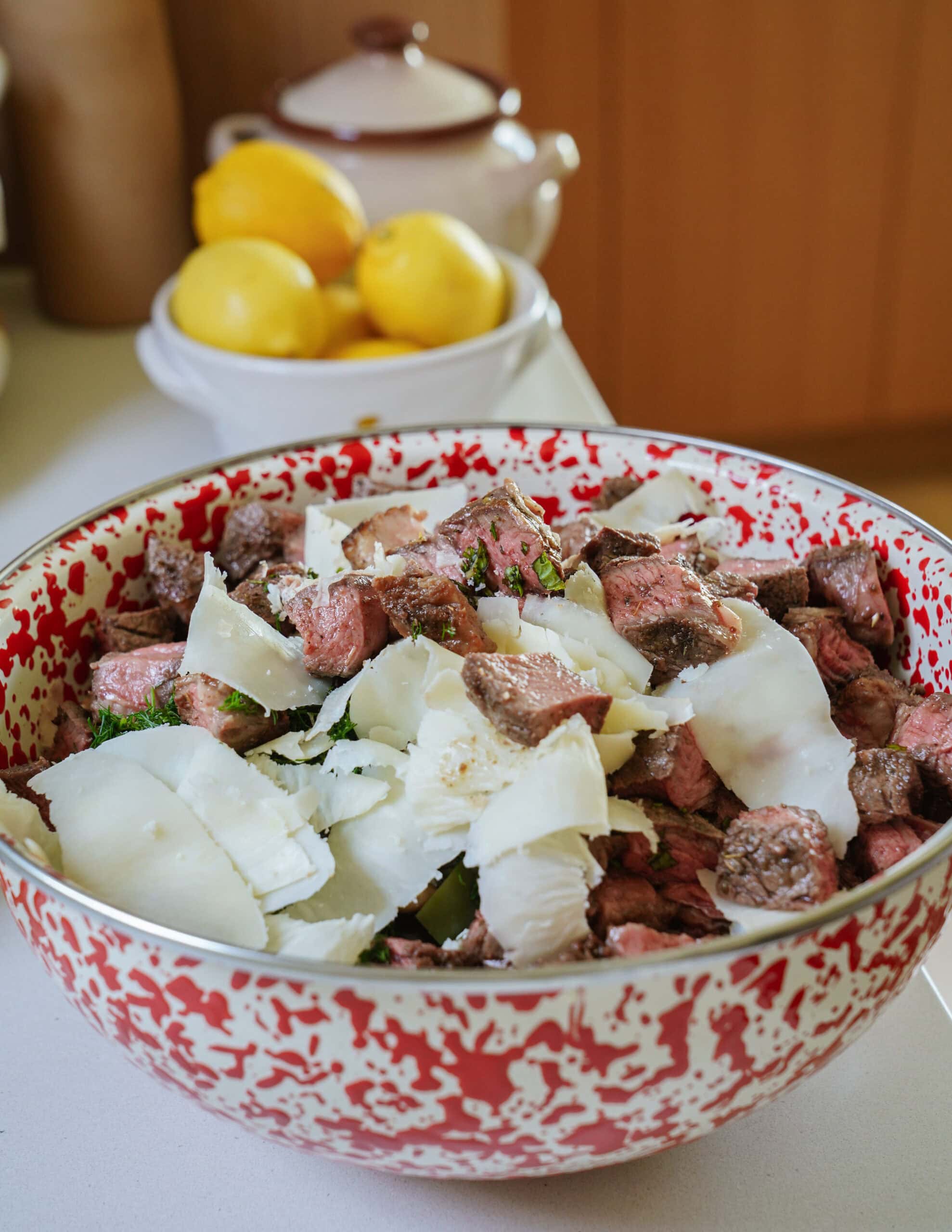 A speckled bowl filled with steak pieces and topped with shaved cheese sits on a kitchen counter. In the background, a white bowl holds several yellow lemons and a ceramic pot with a lid.
