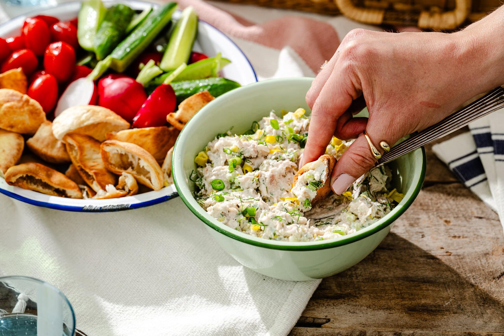 A hand dips pita bread into a bowl of creamy vegetable dip, with a platter of pita chips, tomatoes, cucumbers, and radishes nearby on a wooden table.