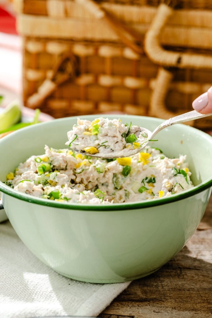 A green bowl filled with creamy chicken salad garnished with chopped green onions and herbs. A spoon is scooping some salad from the bowl. A picnic basket is visible in the background.