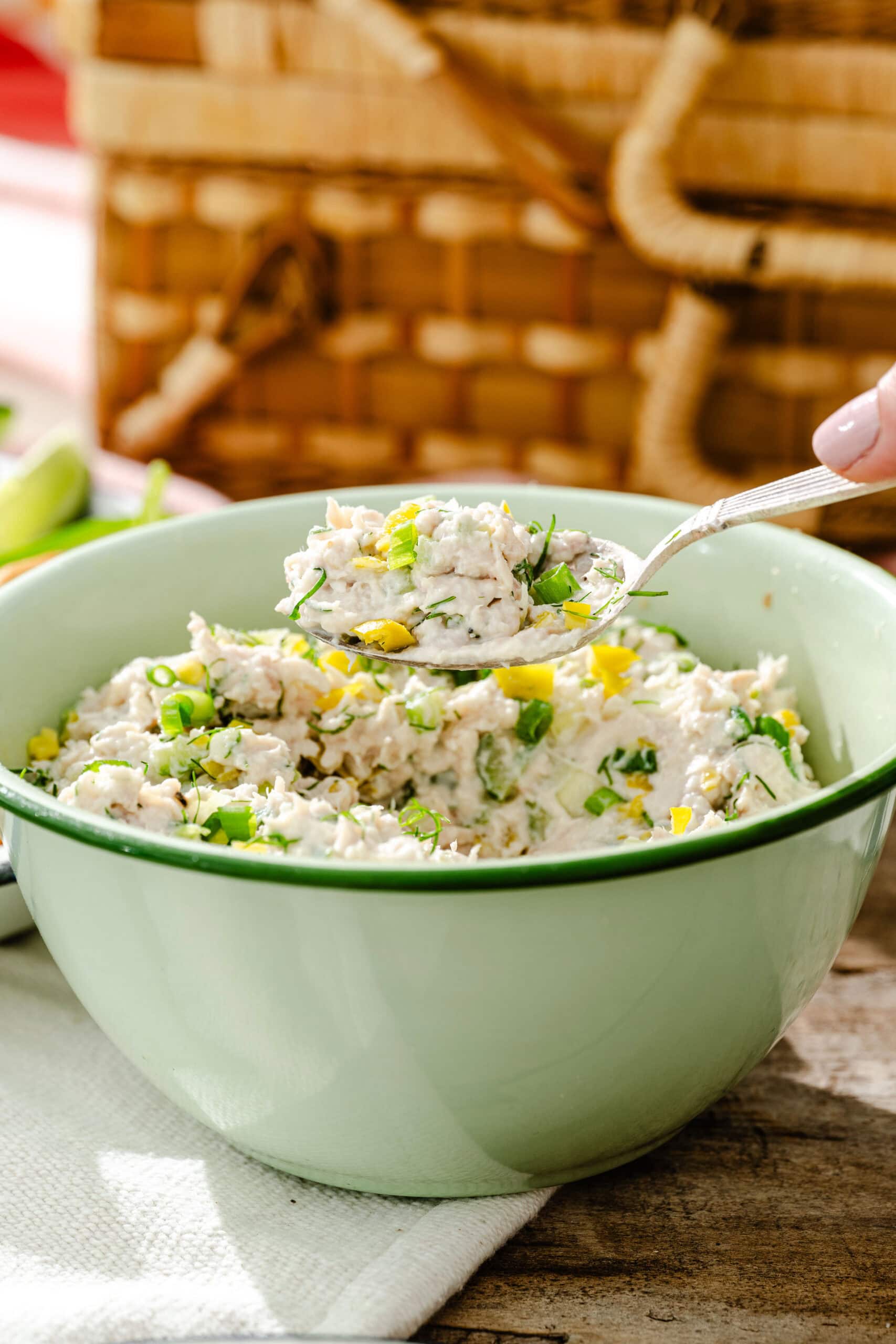 A green bowl filled with creamy chicken salad garnished with chopped green onions and herbs. A spoon is scooping some salad from the bowl. A picnic basket is visible in the background.