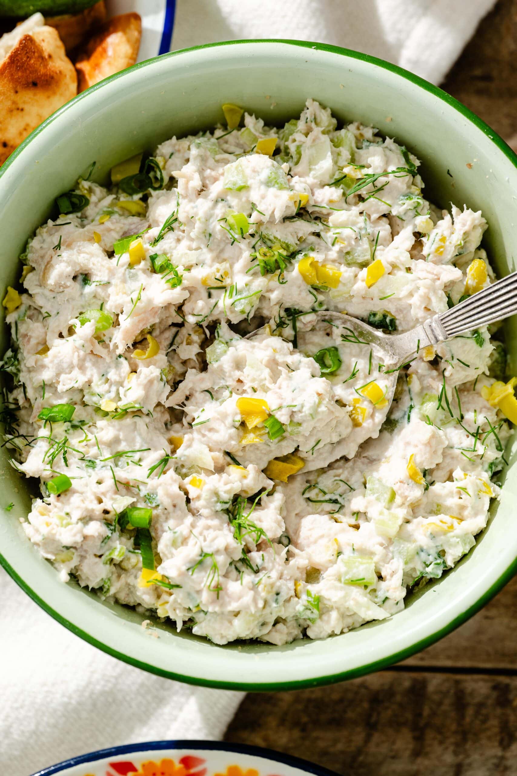 A green bowl filled with a creamy chicken salad mixed with chopped celery, pickles, and fresh herbs, with a spoon resting inside.