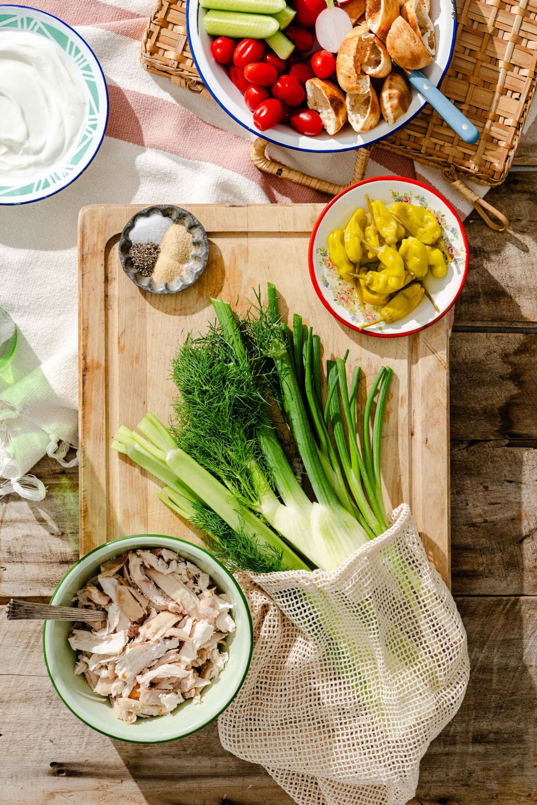 A wooden table set with a cutting board holding fresh dill, green onions, pepperoncinis, and spices. Nearby are a bowl of shredded chicken, pita, cucumber, tomatoes, and a bowl of yogurt dip.