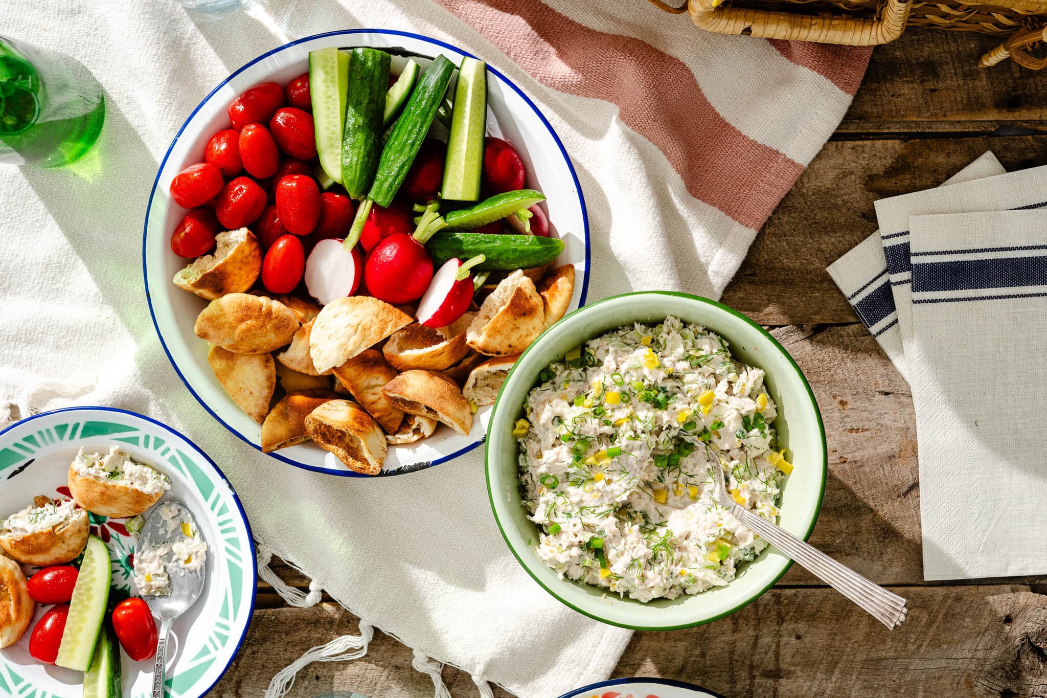 A bowl of pita pieces, cherry tomatoes, okra, and radishes sits next to a bowl of creamy dip garnished with herbs and corn, all on a wooden picnic table with napkins and a blanket.