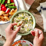 A person scoops creamy chicken salad with herbs from a bowl using pita bread. Fresh vegetables and more pita chips are nearby on a rustic wooden table with cloth napkins.