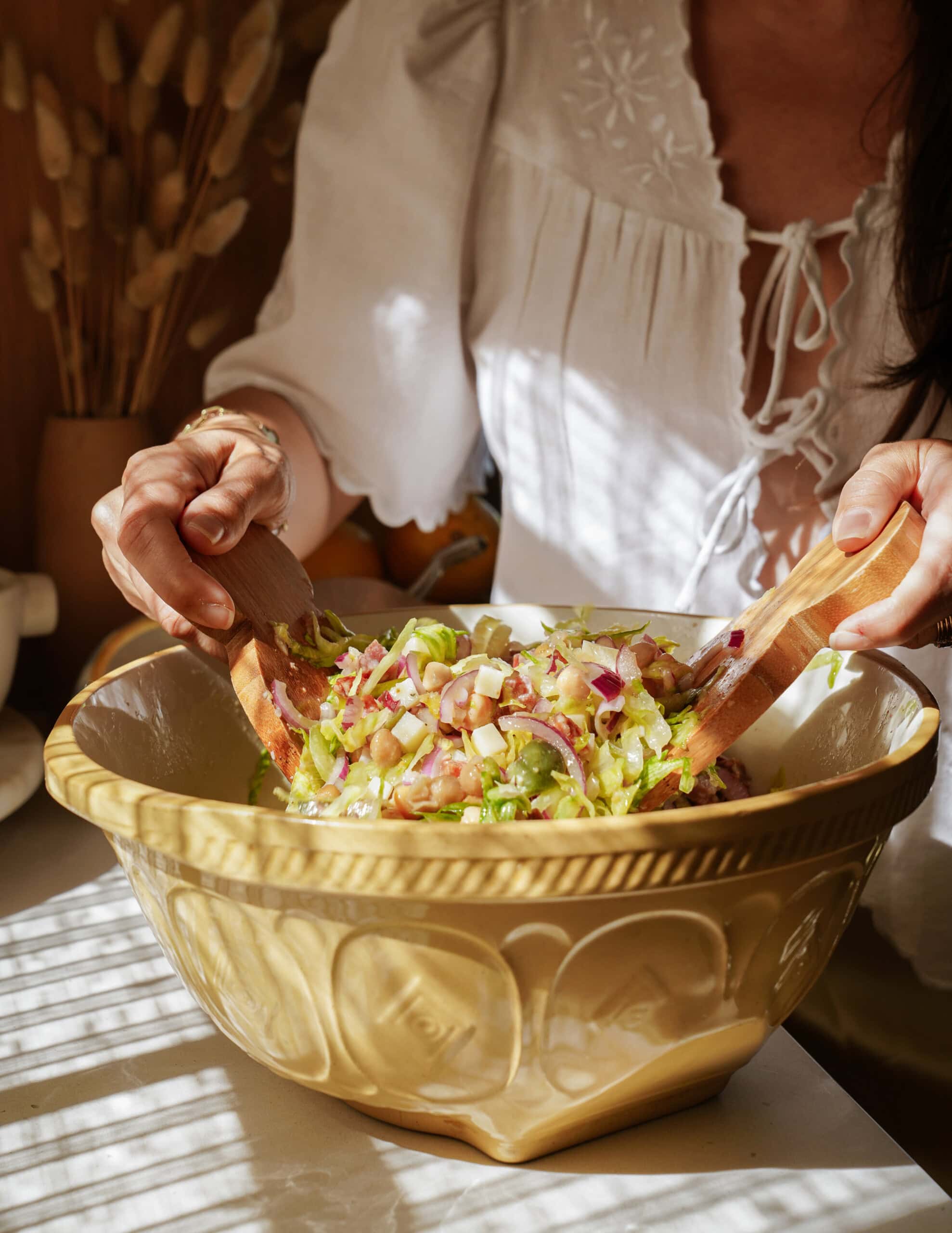 A person in a white blouse uses wooden salad servers to mix a fresh salad with lettuce and red onions in a large yellow ceramic bowl, with sunlight streaming onto the table.