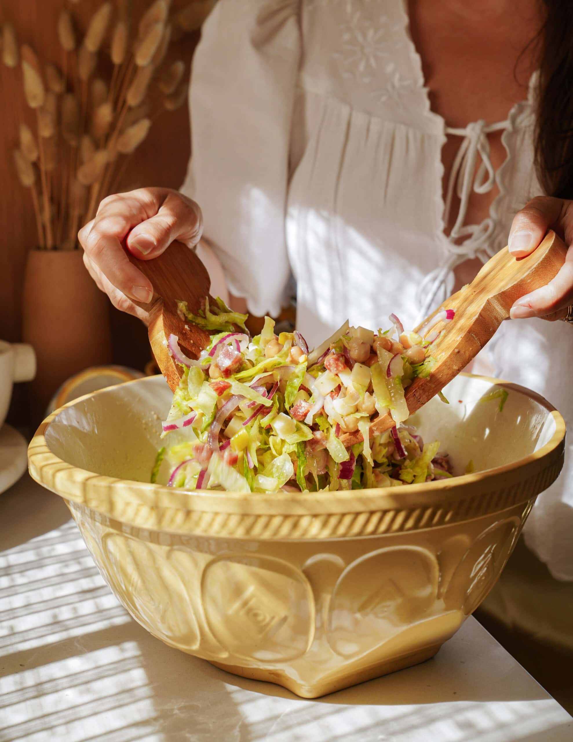 A person in a white blouse uses wooden salad tongs to mix a fresh salad with lettuce and red onions in a large yellow ceramic bowl on a sunlit table.