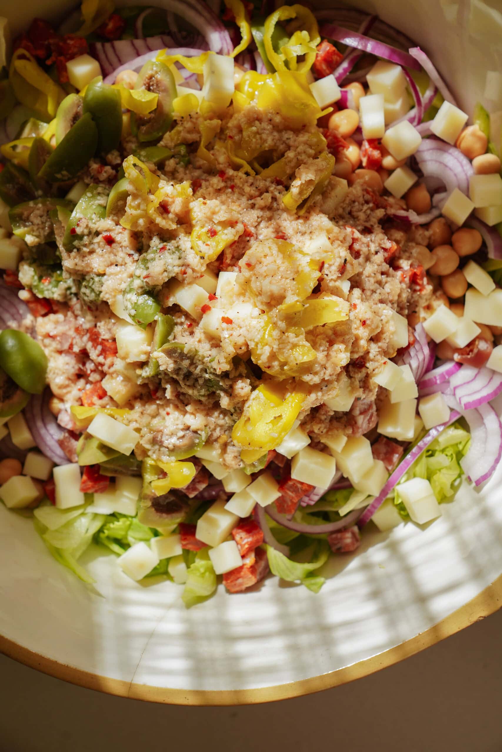 A close-up of a salad bowl with layers of sliced red onions, chickpeas, chopped lettuce, diced cheese, green olives, pepperoncini peppers, red bell peppers, and a tuna mixture on top.
