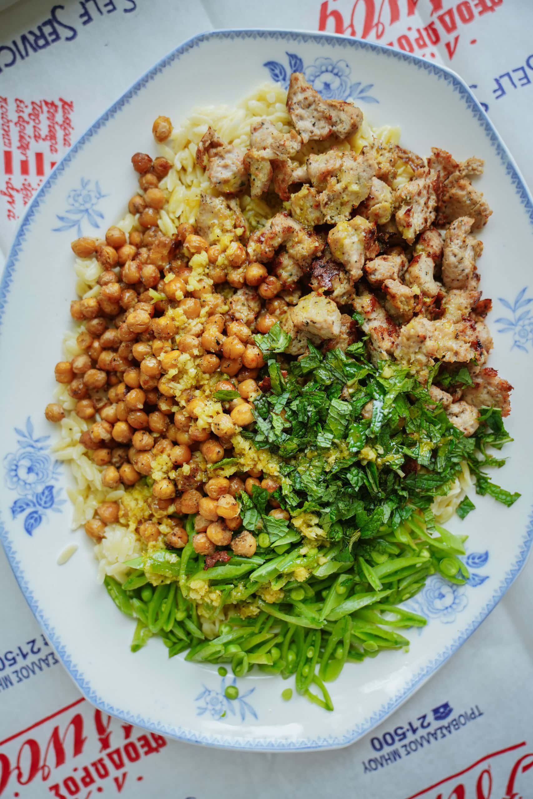 A plate with orzo pasta, roasted chickpeas, grilled chicken pieces, chopped fresh herbs, and green beans, arranged in separate sections on a white and blue floral dish.