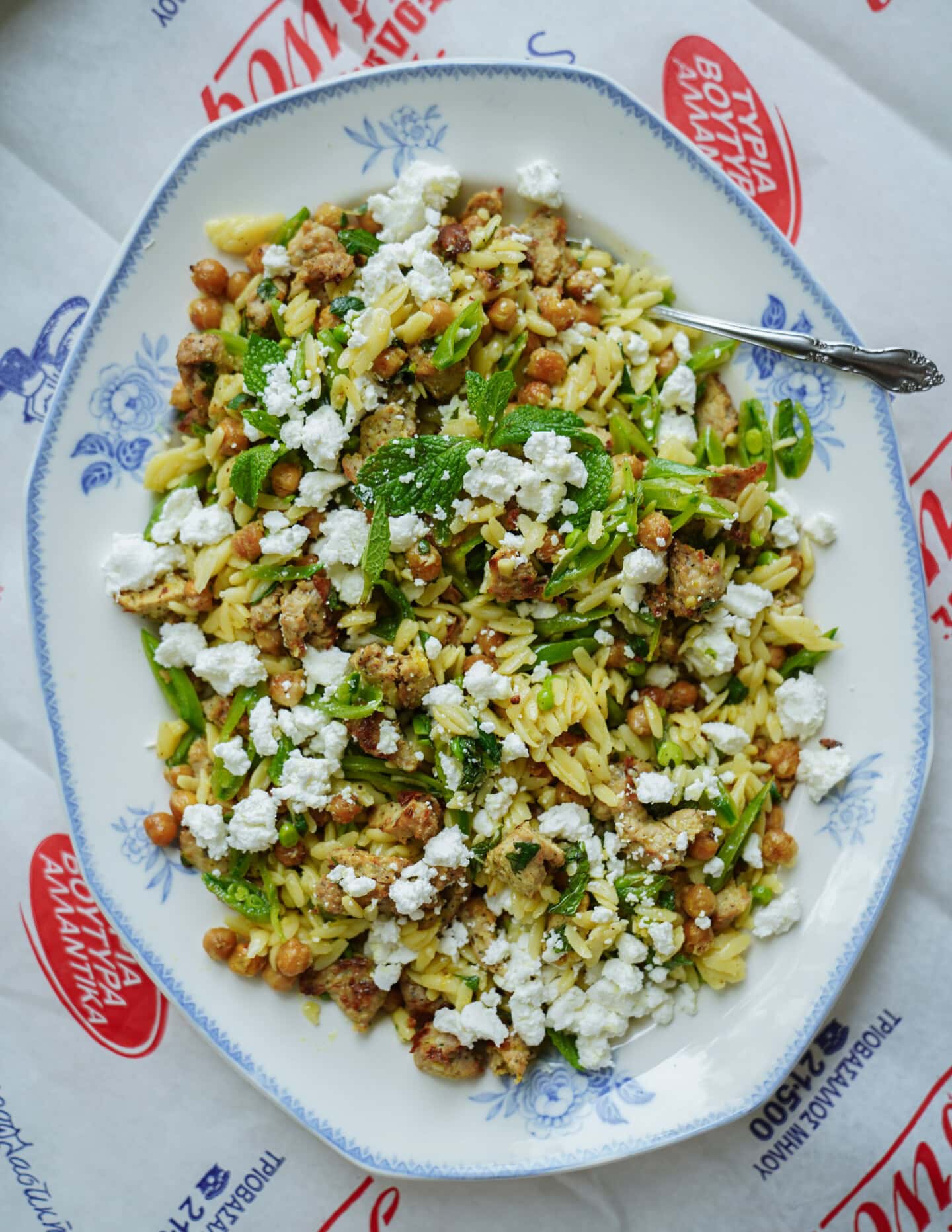 An oval platter of orzo pasta salad with chickpeas, crumbled feta cheese, green herbs, and sausage pieces, served on a patterned blue and white plate with a spoon on the side.
