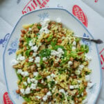 A platter of orzo salad with crumbled feta cheese, chickpeas, fresh mint, and herbs, garnished with greens and served with a spoon on a patterned white plate.