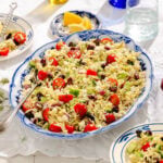 A large blue and white platter filled with orzo pasta salad featuring cherry tomatoes, cucumber, kalamata olives, red onion, feta cheese, and fresh herbs. Lemon wedges and plates of salad are on a white tablecloth nearby.
