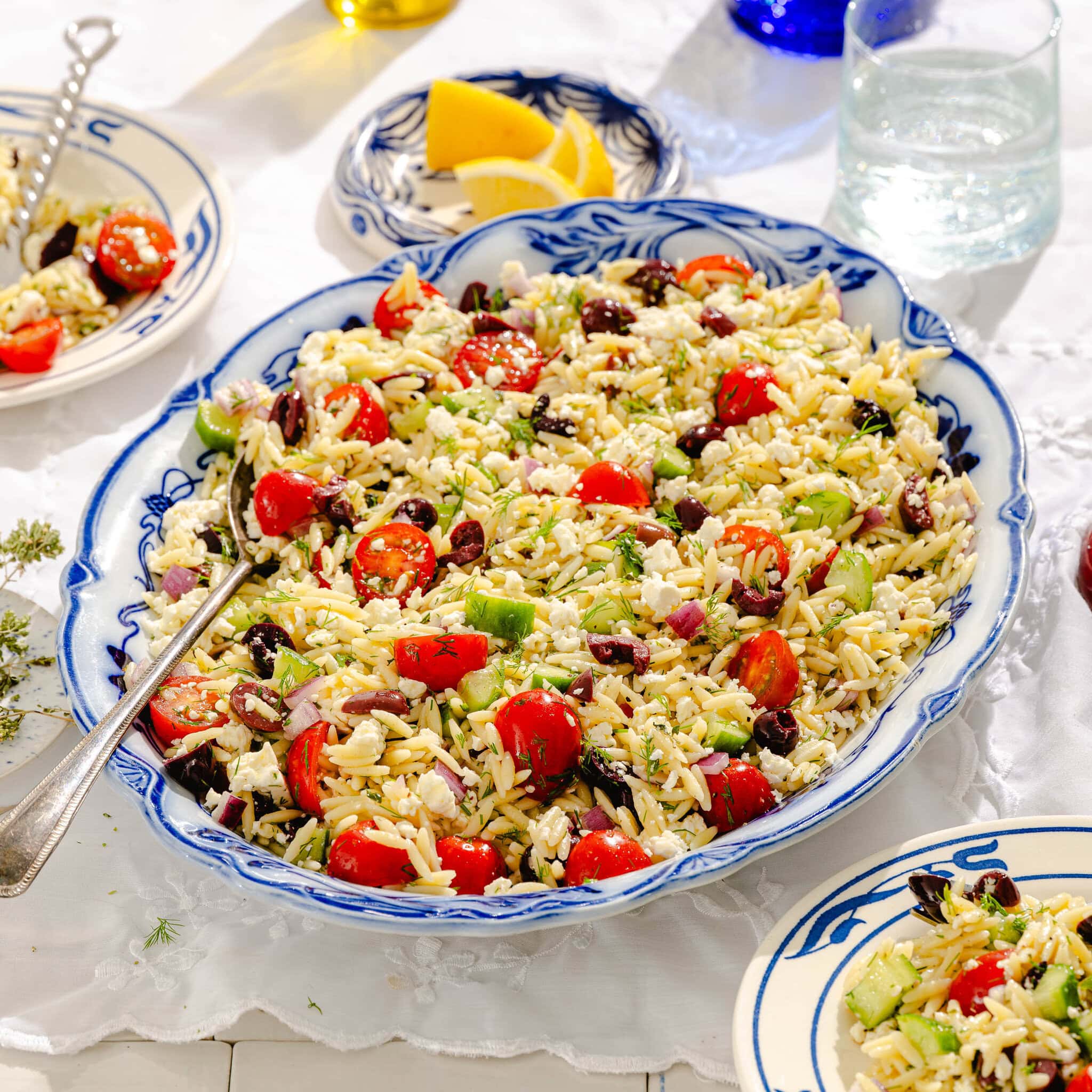 A large blue and white platter filled with orzo pasta salad featuring cherry tomatoes, cucumber, kalamata olives, red onion, feta cheese, and fresh herbs. Lemon wedges and plates of salad are on a white tablecloth nearby.