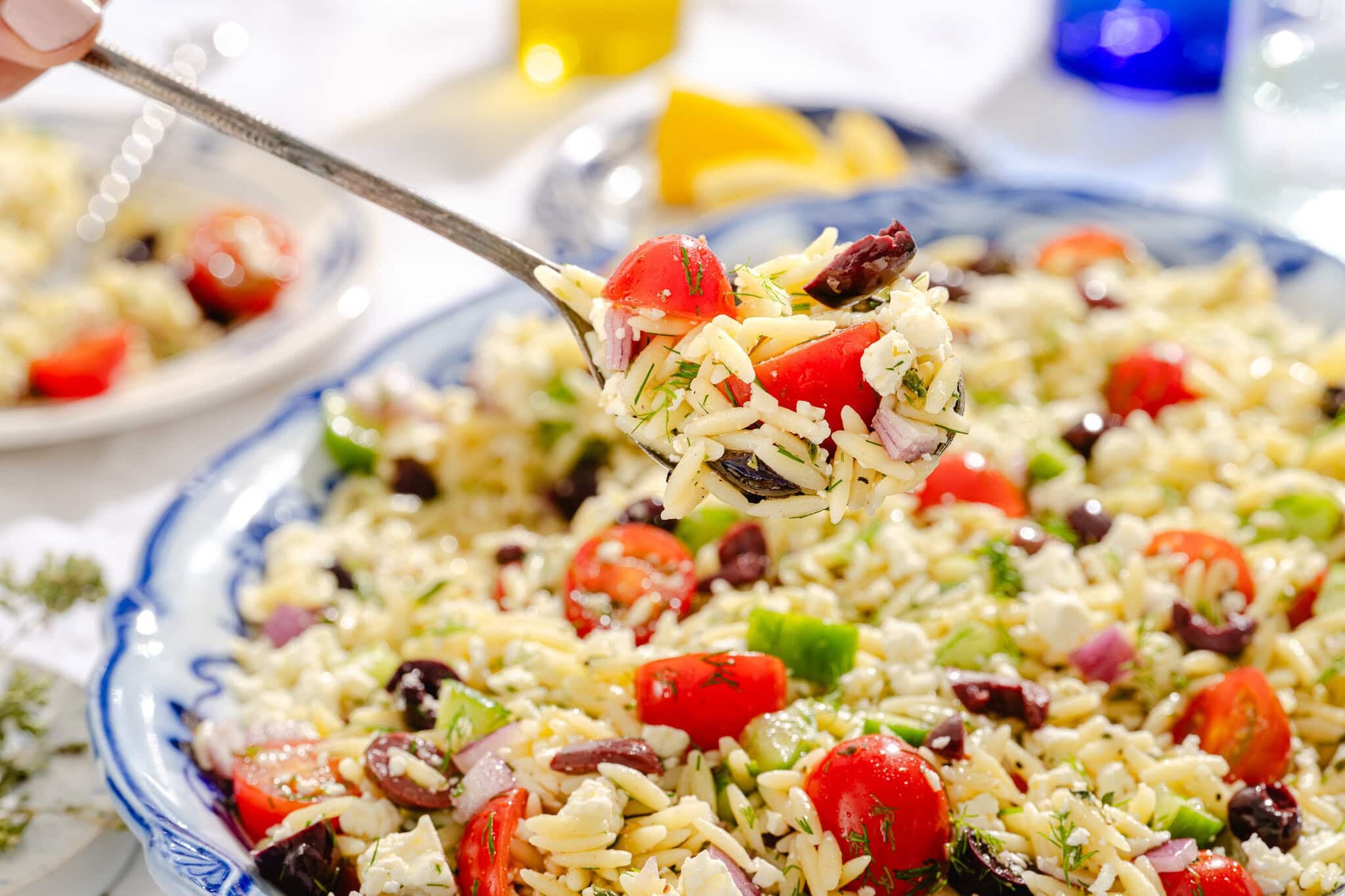A close-up of a spoonful of orzo salad with cherry tomatoes, olives, diced red onion, green pepper, feta cheese, and fresh herbs, taken from a large blue plate of the same salad in bright, natural light.