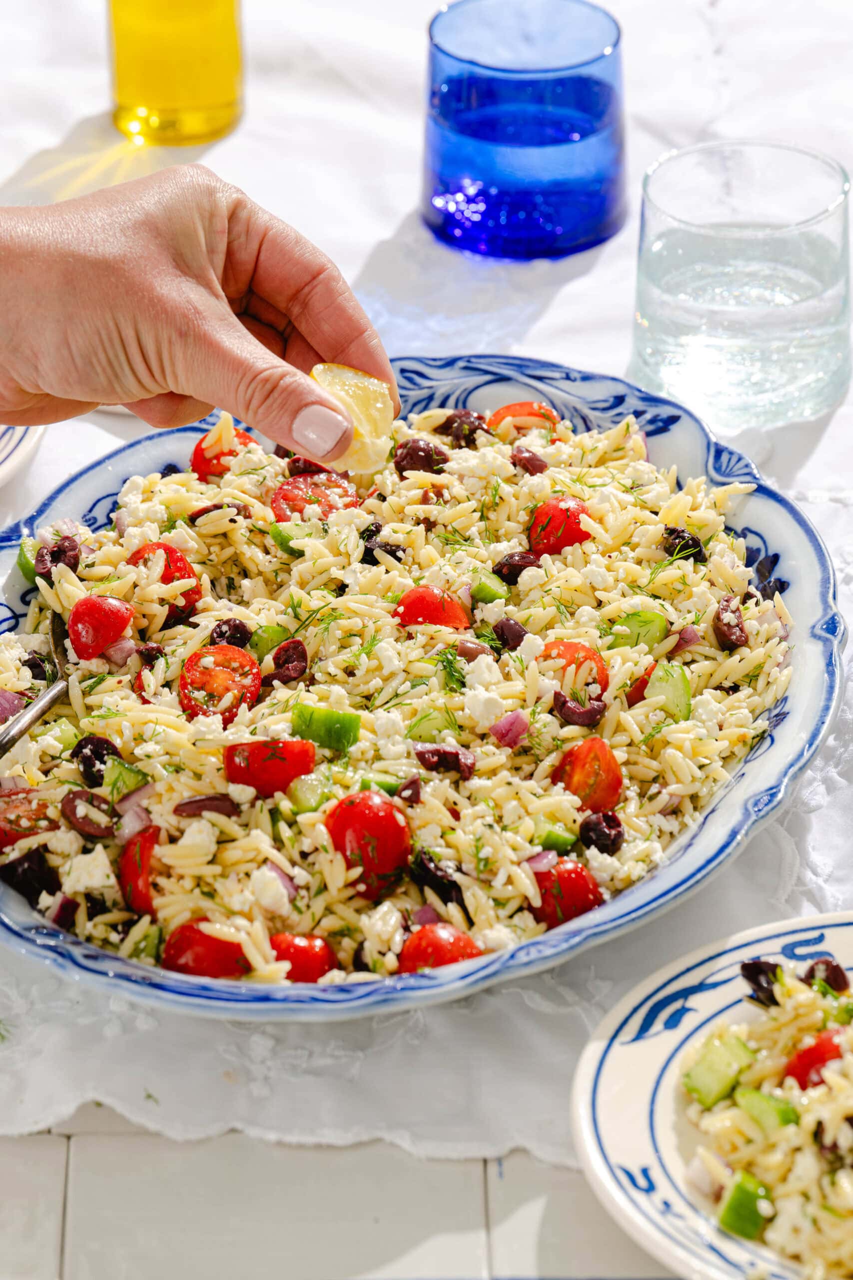 A hand squeezes a lemon wedge over a large bowl of orzo salad with cherry tomatoes, cucumber, olives, and feta cheese. Two glasses and an olive oil bottle are in the background, and a small plate of salad sits nearby.