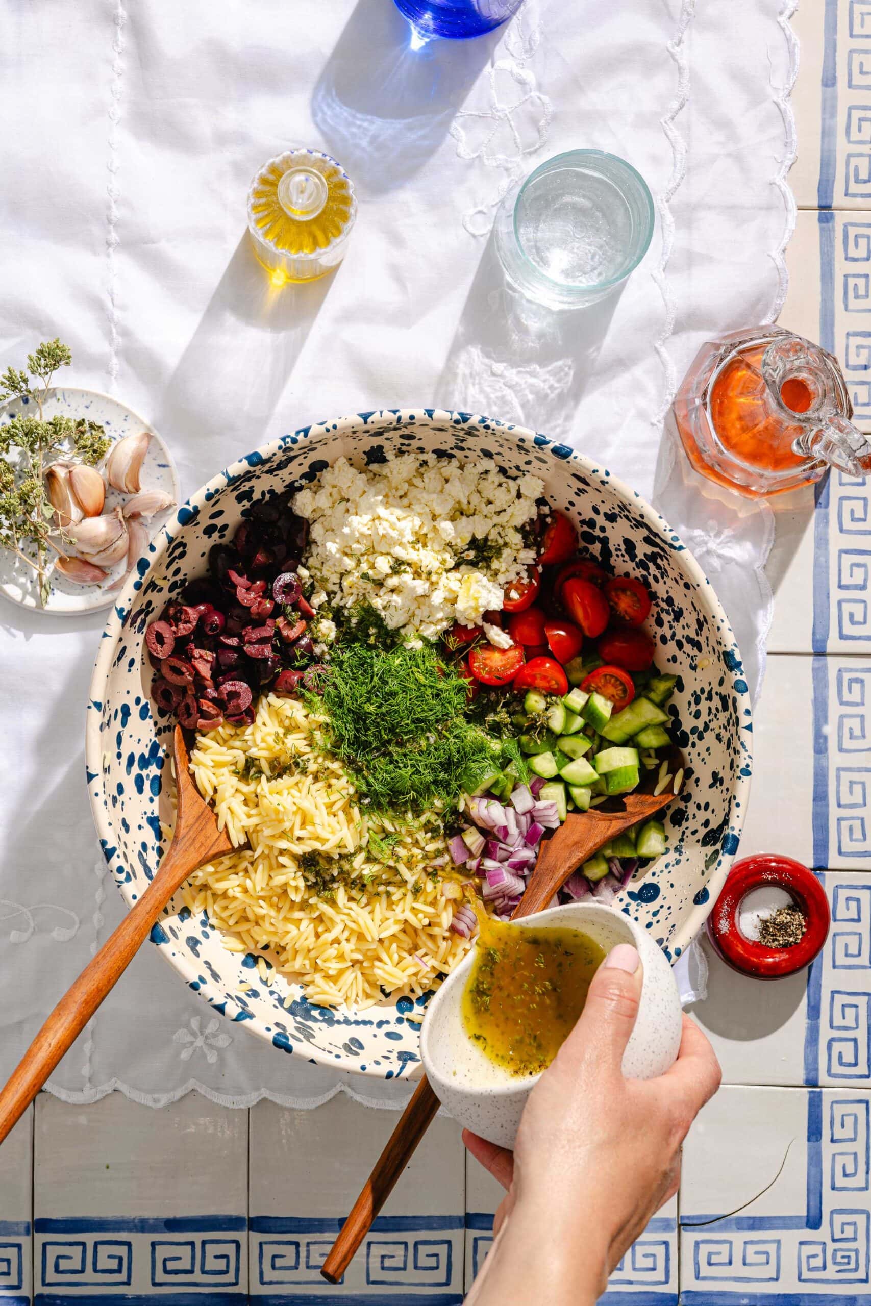 A hand pours dressing over a large bowl of orzo salad with feta, olives, cherry tomatoes, cucumber, red onion, and fresh herbs. The bowl sits on a white tablecloth with drinks, olive oil, and garlic nearby.
