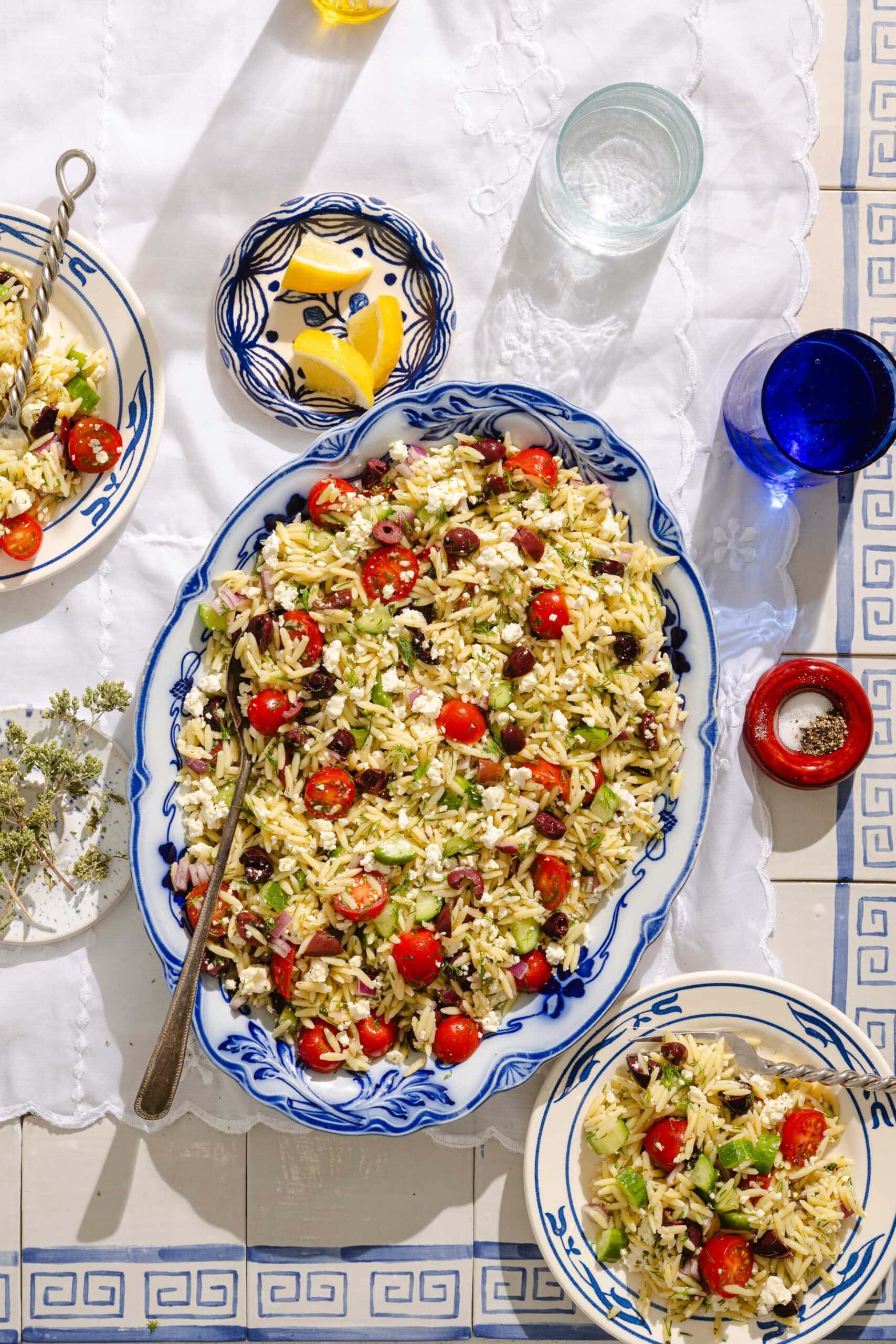 A large blue and white platter filled with orzo salad featuring cherry tomatoes, olives, feta, and herbs sits on a patterned tablecloth, surrounded by plates, glasses, and a small dish with lemon wedges.