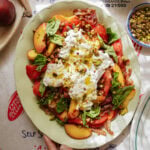 A colorful salad on a platter with tomatoes, peach slices, burrata cheese, fresh basil, crushed pistachios, and olive oil, arranged on a table with a Greek-themed tablecloth.