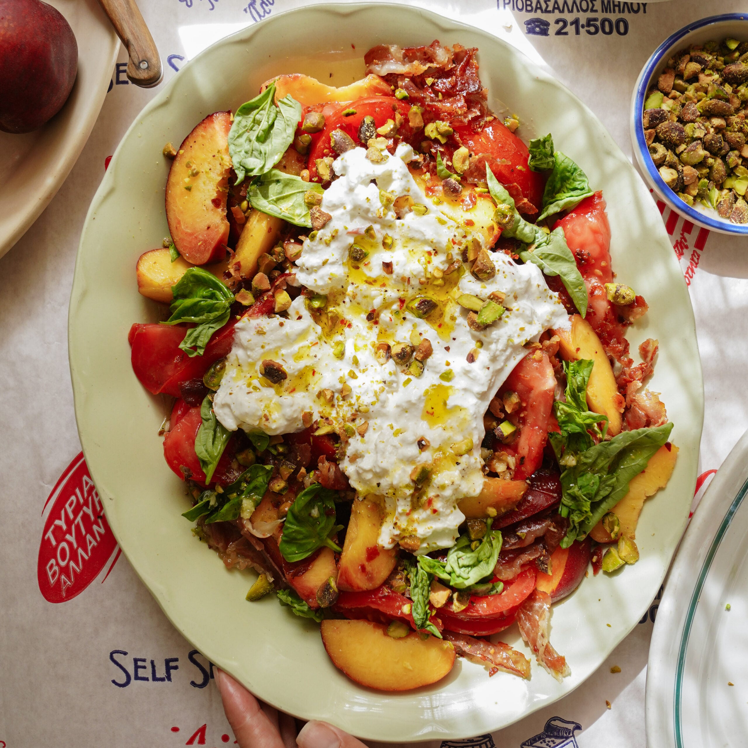 A colorful salad on a platter with tomatoes, peach slices, burrata cheese, fresh basil, crushed pistachios, and olive oil, arranged on a table with a Greek-themed tablecloth.
