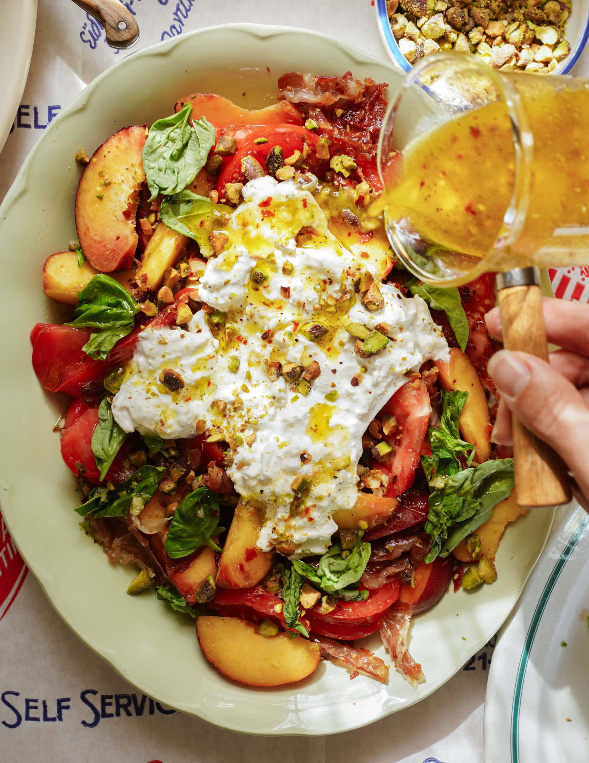 A hand pours vinaigrette over a salad with burrata, sliced peaches, tomatoes, fresh basil, and chopped pistachios in a bowl on a table.