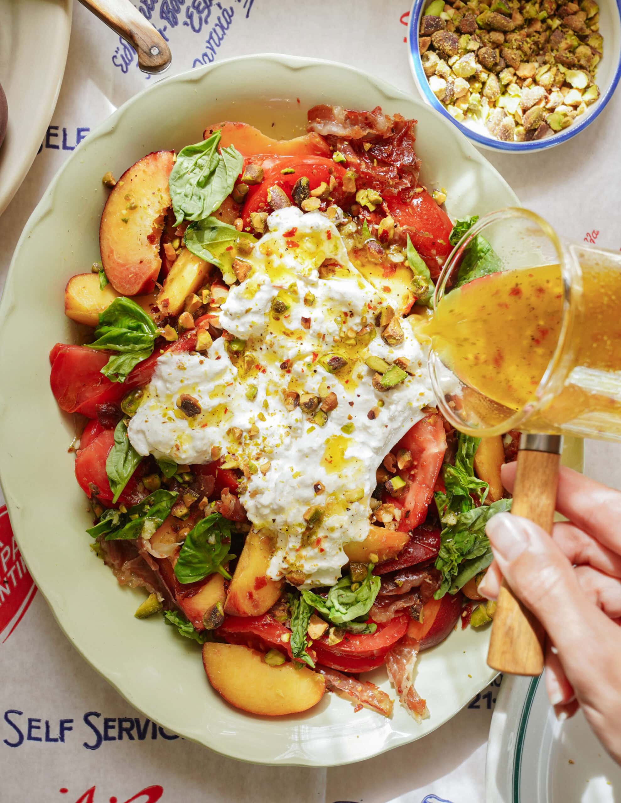 A hand pours dressing onto a colorful salad with burrata cheese, peach slices, tomatoes, spinach, and crushed pistachios on a platter. A bowl of pistachios is nearby on a table with a printed tablecloth.