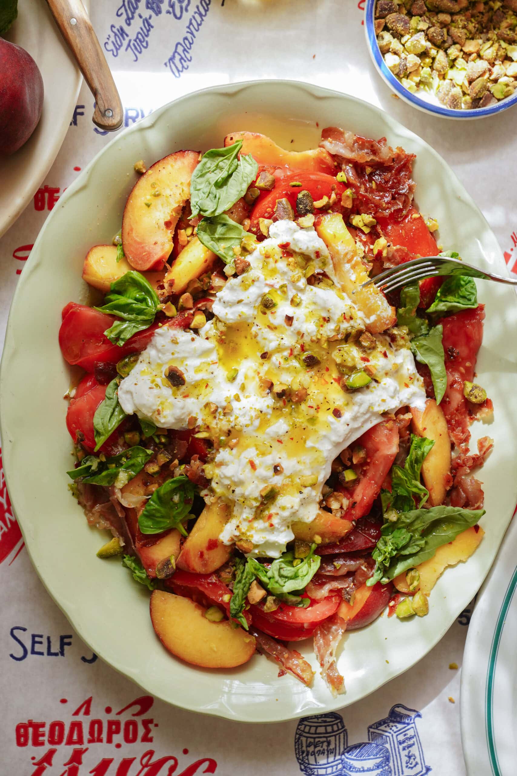A plate of salad with sliced peaches, tomatoes, fresh basil, burrata cheese, chopped pistachios, and a drizzle of olive oil sits on a patterned tablecloth. A fork rests on the plate.