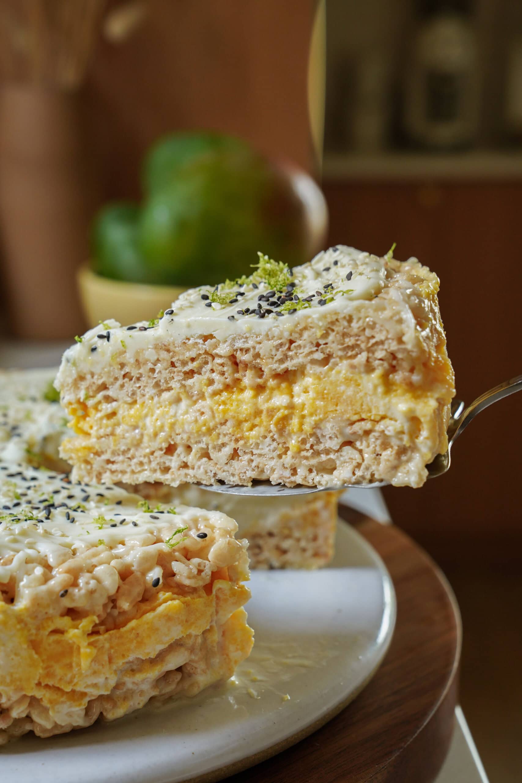 A close-up of a slice of creamy layered dessert, possibly with puffed rice and yellow filling, being lifted from a larger cake on a plate. The dessert is topped with white frosting and sprinkled with black and green bits.