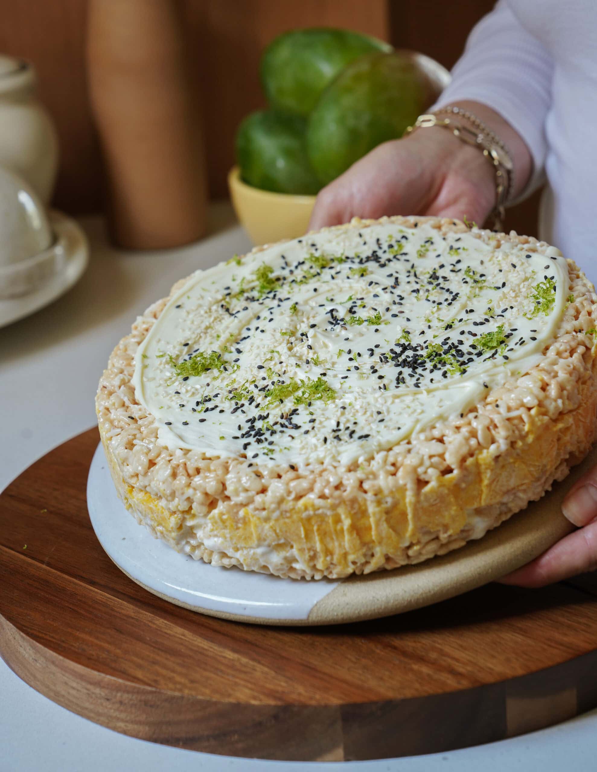 A person holds a round cake with white frosting, topped with green zest and black seeds. The cake’s sides are covered in crispy rice cereal. Mangos and kitchen items are visible in the background.