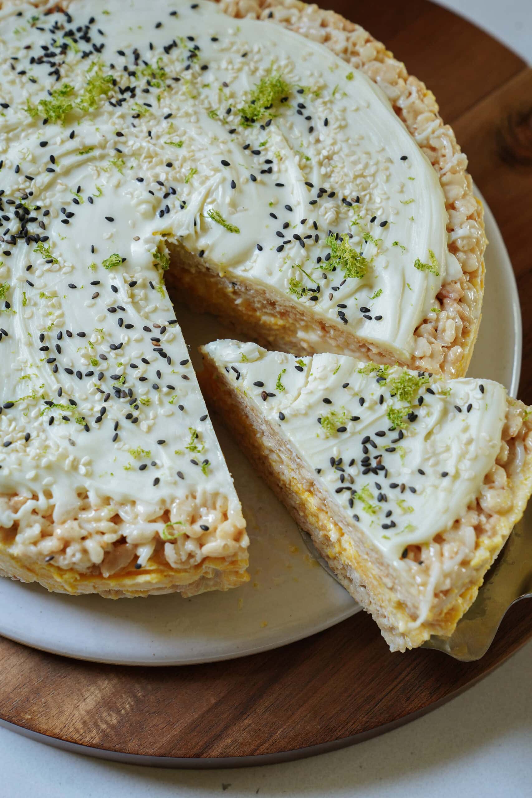 A creamy, round cake topped with white frosting, black sesame seeds, and lime zest sits on a plate. A single slice is being removed, revealing a layered, rice cereal-like texture inside.