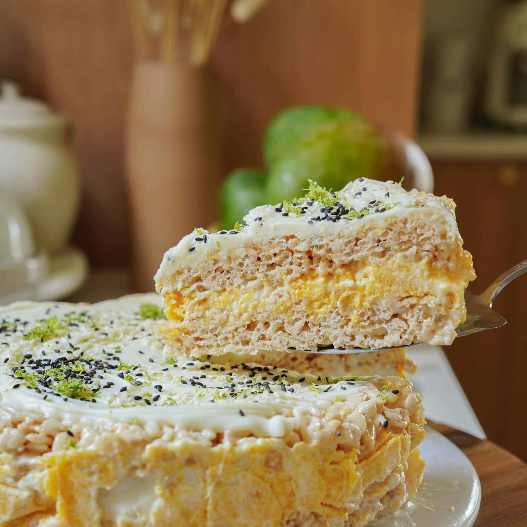 A close-up of a creamy layered cake with a slice being lifted, showing textures of cream and sponge. The cake is topped with white icing, green sprinkles, and black seeds. A blurred background features kitchen items and fruit.