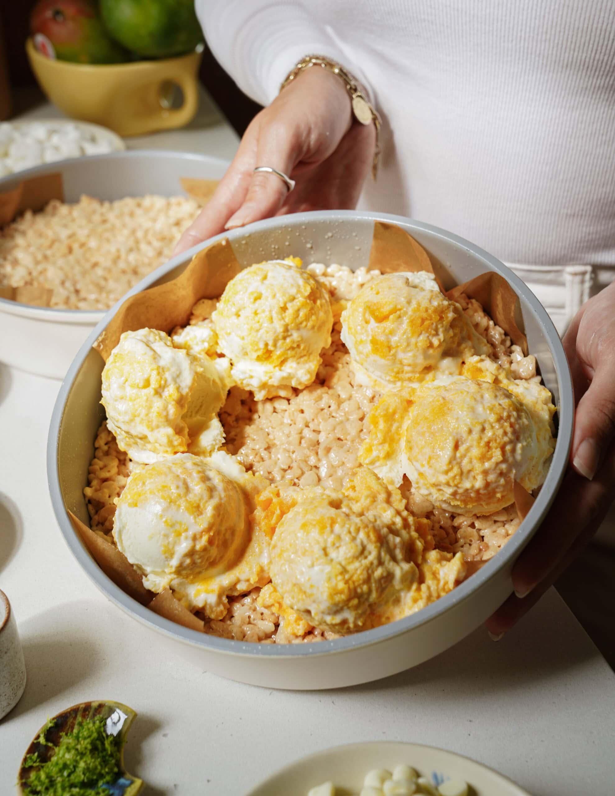 A person holding a round pan filled with rice crispy treats, topped with six large scoops of what appears to be vanilla and orange swirled ice cream or frosting. Other baking ingredients are visible in the background.