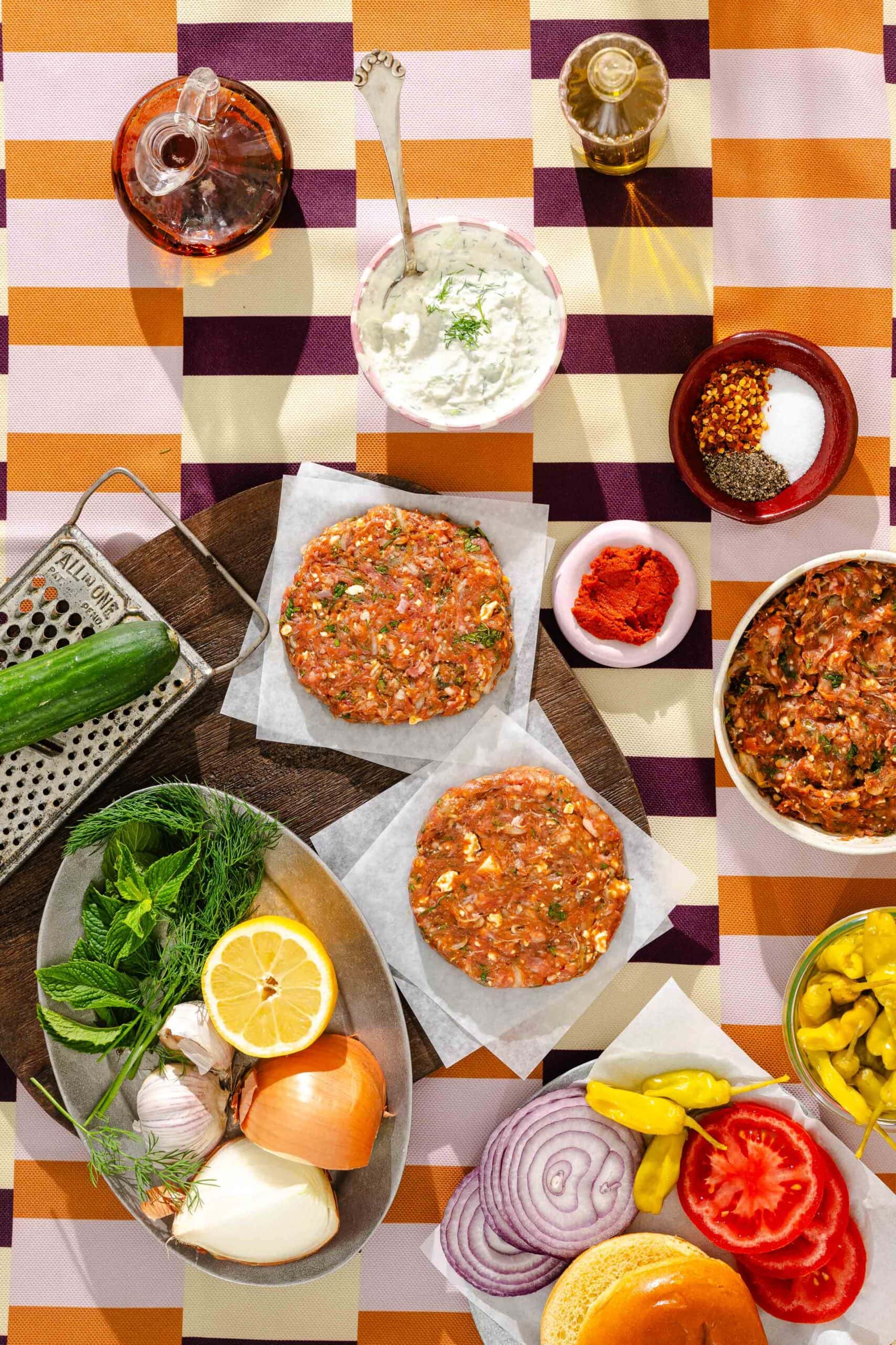 Overhead view of ingredients for making burgers, including raw burger patties, sliced vegetables, spices, lemon, fresh herbs, grated cucumber, sauce in a bowl, and a burger bun on a colorful striped tablecloth.