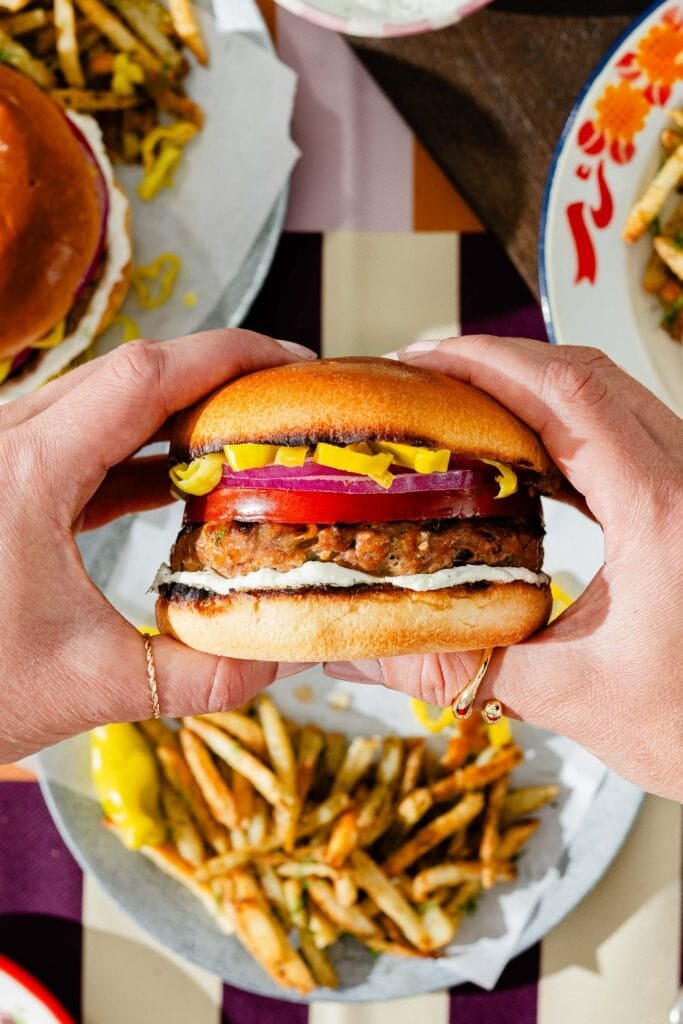A person holds a hamburger with pickles, red onion, and sauce in both hands, with plates of seasoned fries and another burger visible in the background on a checkered tablecloth.