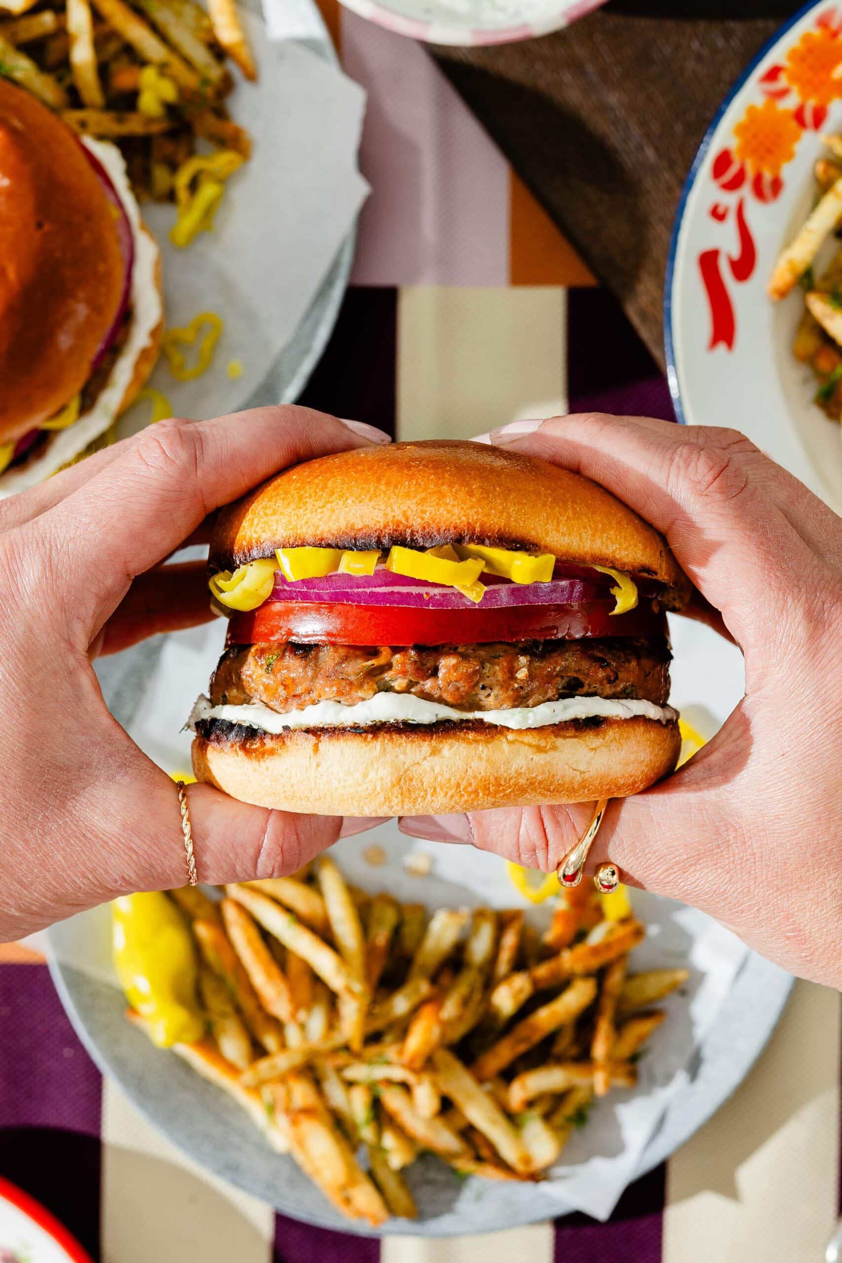 A person holds a hamburger with pickles, red onion, and sauce in both hands, with plates of seasoned fries and another burger visible in the background on a checkered tablecloth.