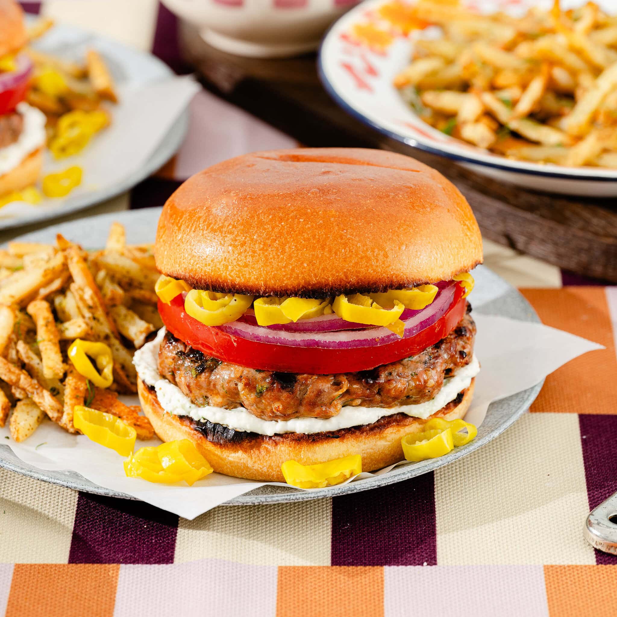 A grilled burger with tomato, red onion, sliced yellow peppers, and a creamy spread in a brioche bun, served with seasoned fries on a plate on a checkered tablecloth.