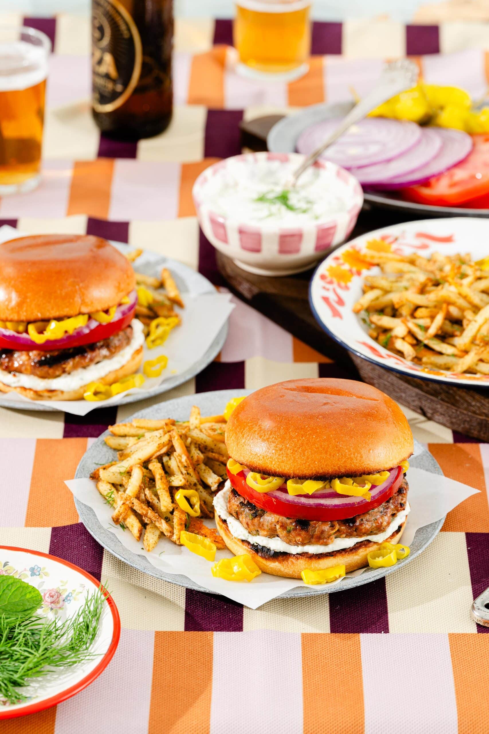 Two burgers with red onion, sliced peppers, and lettuce are served on plates with seasoned fries. On the table are bowls of sliced onions, tomatoes, dip, and glasses of beer, all set on a colorful checkered tablecloth.