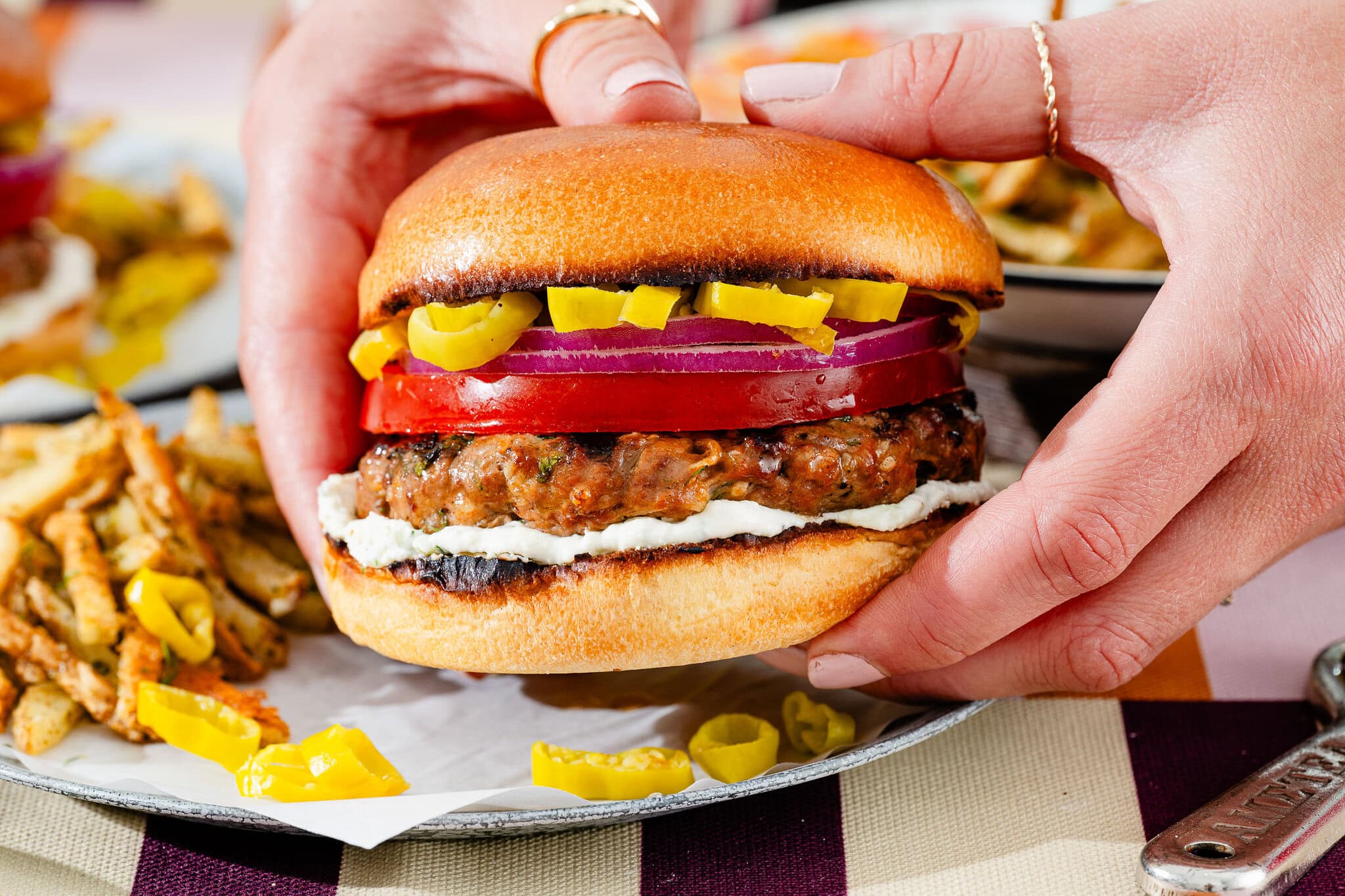 Close-up of hands holding a hamburger with a beef patty, sliced tomatoes, red onion, banana peppers, and a spread, with fries and more banana peppers on a plate in the background.
