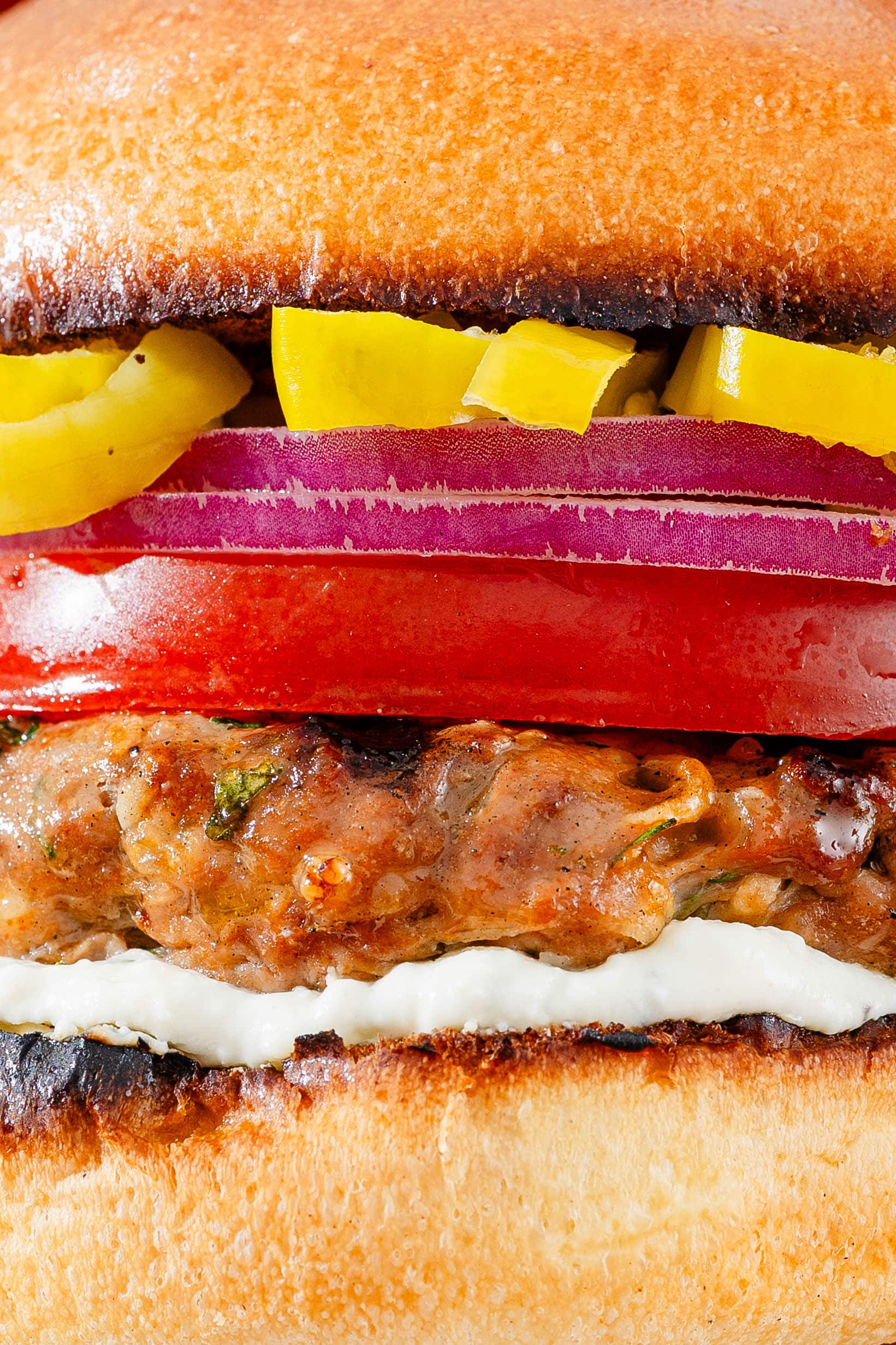 Close-up of a burger showing layers of sliced yellow peppers, red onions, tomato, a grilled beef patty, white sauce, and a toasted bun.