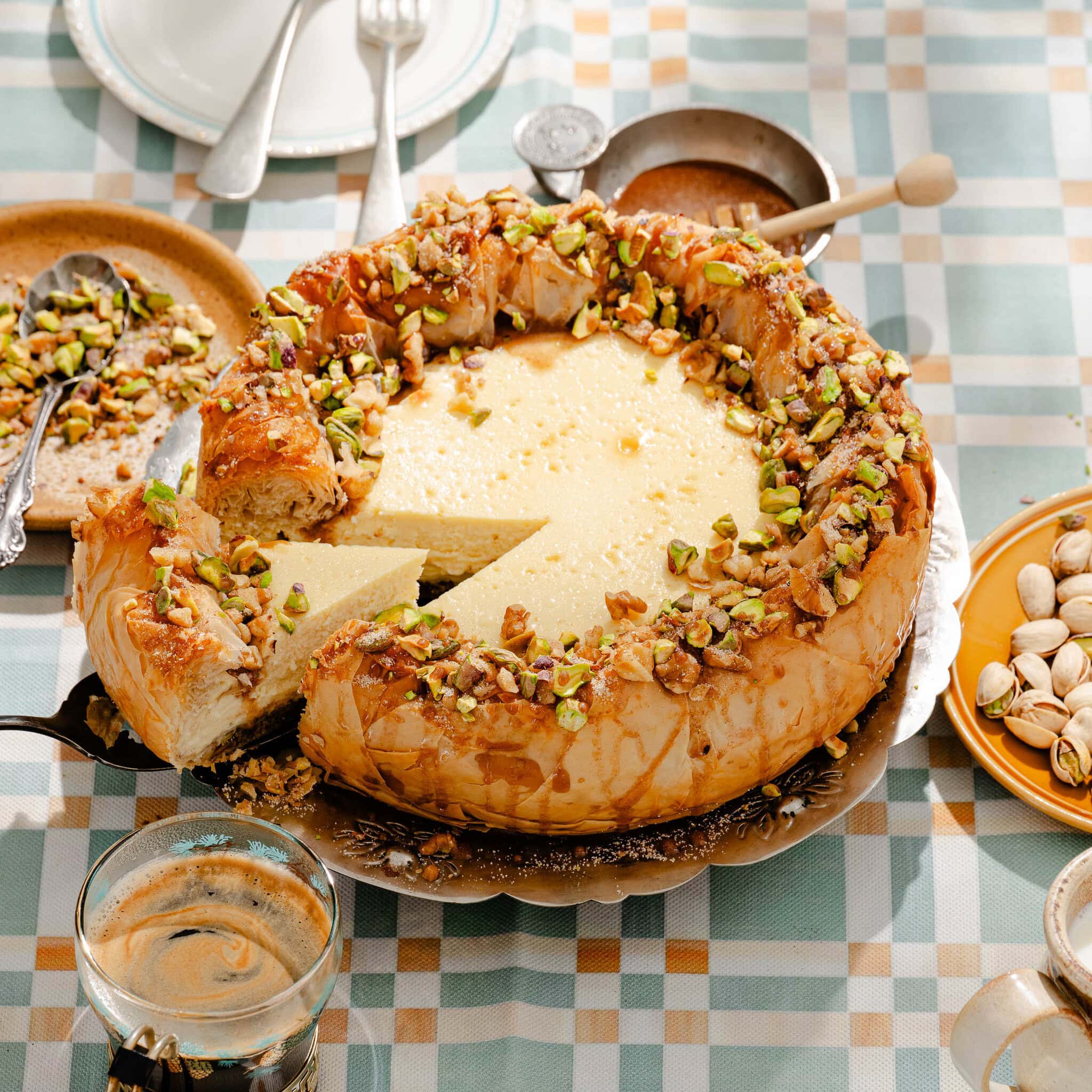 A creamy cheesecake with a flaky crust, topped with chopped pistachios and syrup, sits on a checkered tablecloth. A slice has been cut and served on a plate nearby. Glasses and utensils are also visible.