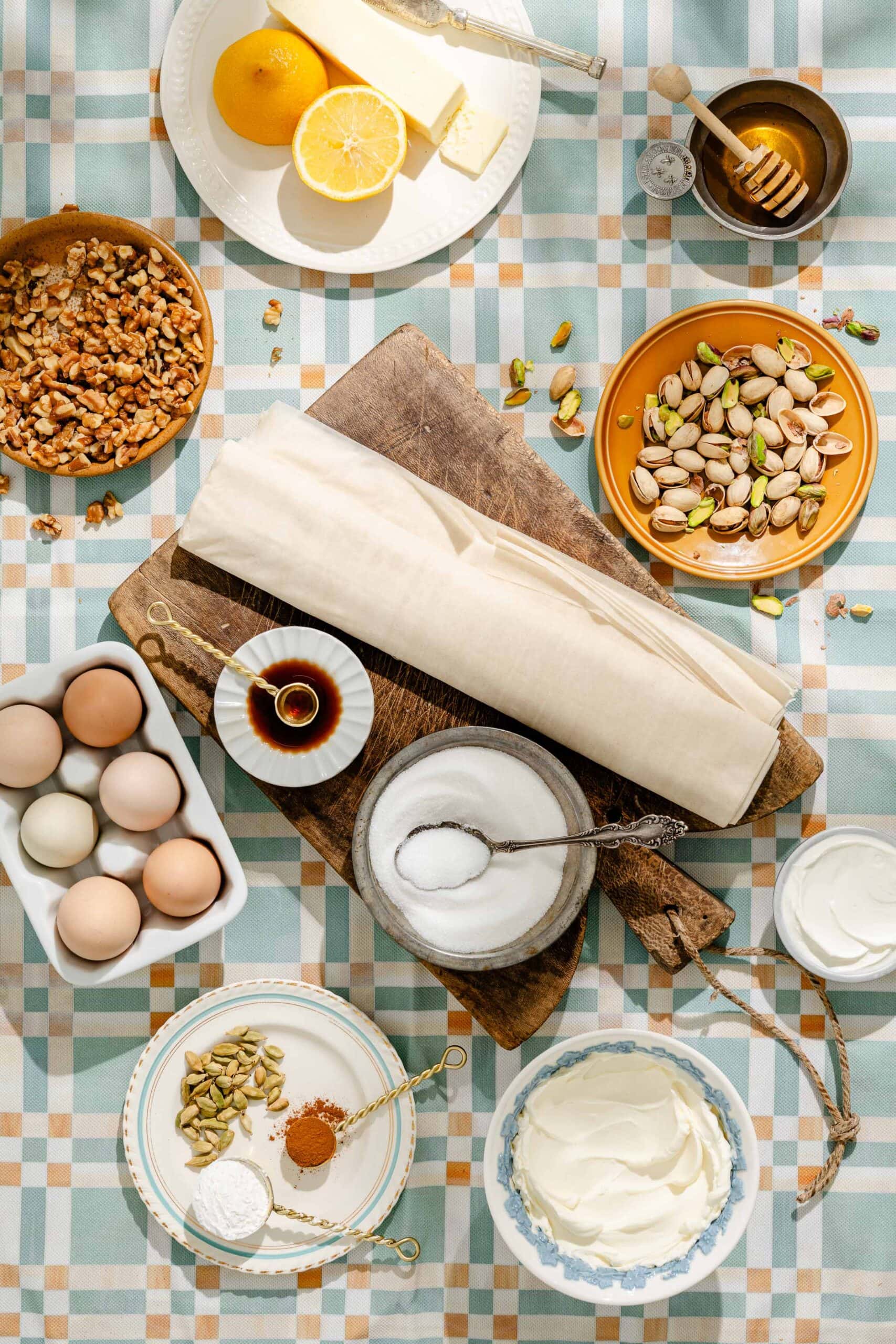 Top-down view of ingredients on a checkered tablecloth, including eggs, nuts, lemon, honey, vanilla, sugar, spices, pastry sheets, and a bowl of cream. Items are arranged neatly around a wooden cutting board.