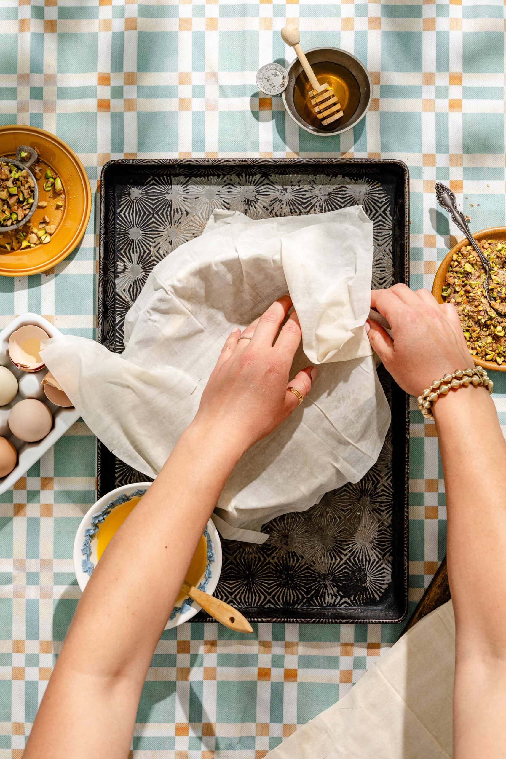 A person prepares pastry on a baking tray lined with parchment, surrounded by bowls of eggs, pistachios, honey, and melted butter on a patterned tablecloth.