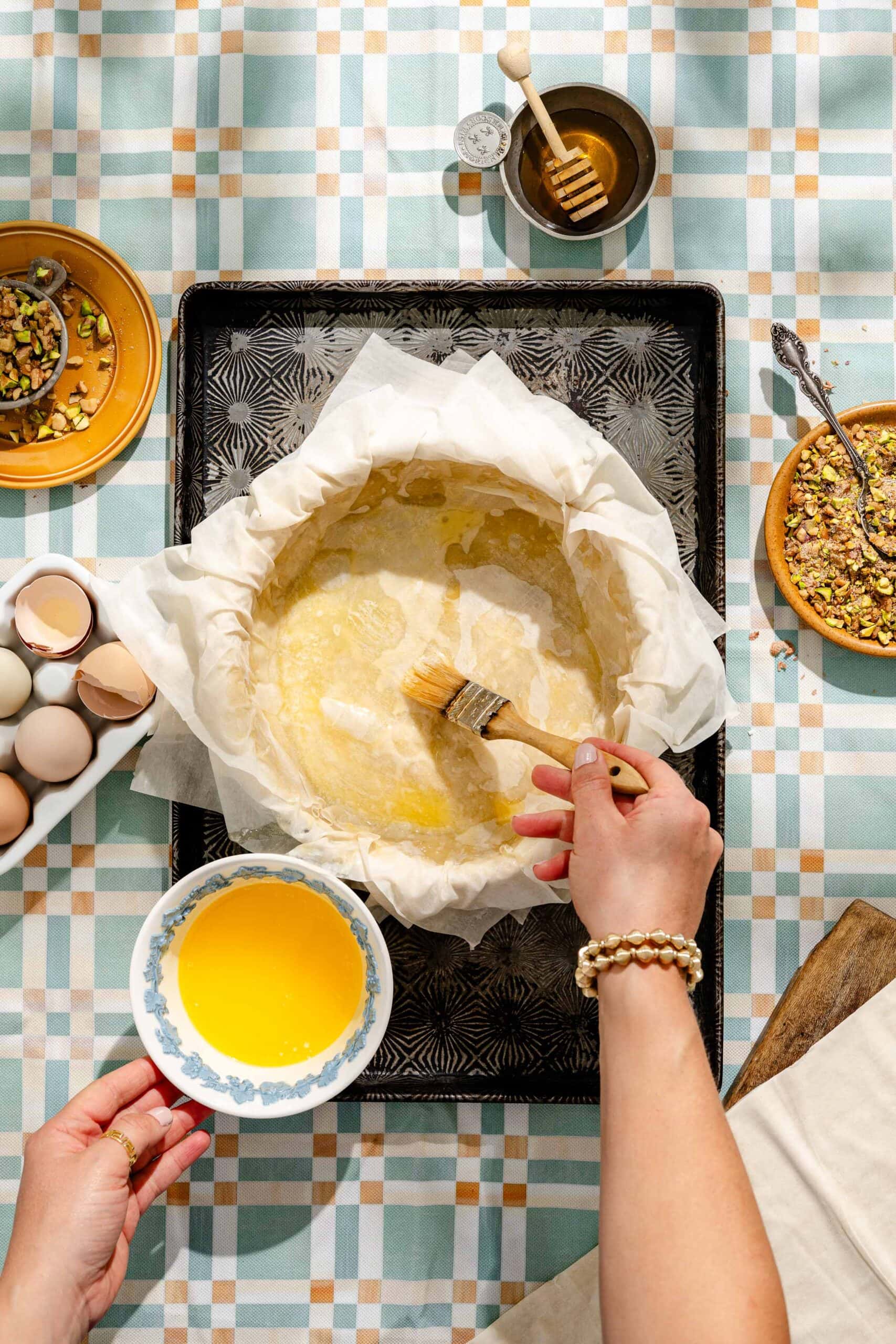 Hands brushing melted butter onto a round pastry in a parchment-lined pan; eggs, nuts, honey, and a patterned tablecloth are visible around the workspace.