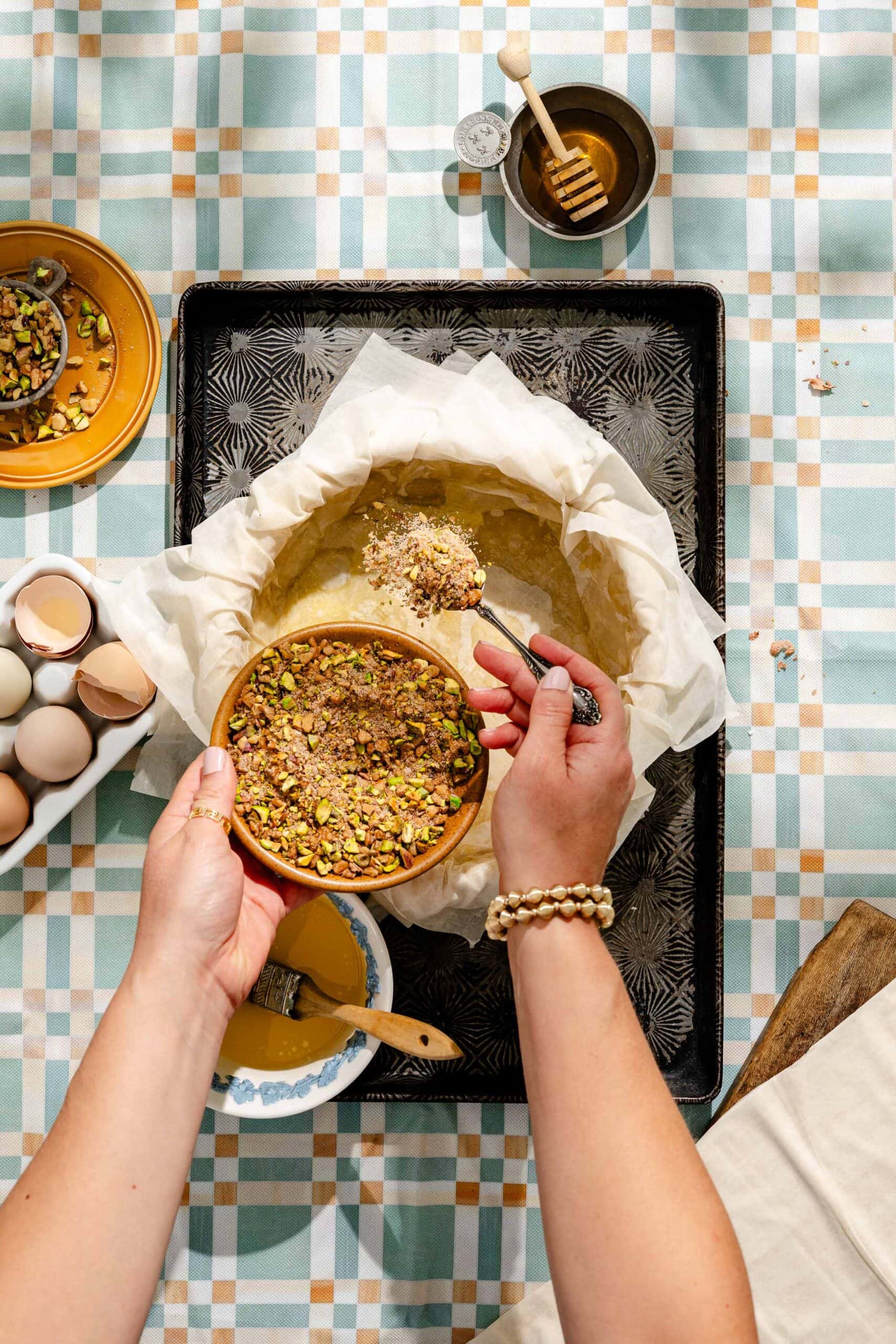 A person sprinkles chopped pistachios and nuts from a bowl into a parchment-lined baking pan on a tray, surrounded by eggs, honey, and other baking ingredients on a patterned tablecloth.